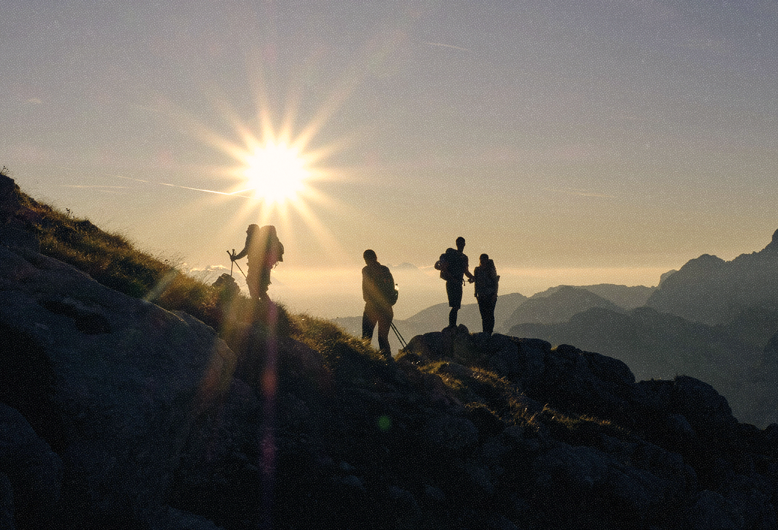 Four hikers with backpacks standing and sitting on rocky terrain at sunset, mountains in the background, sunlight creating a starburst effect.