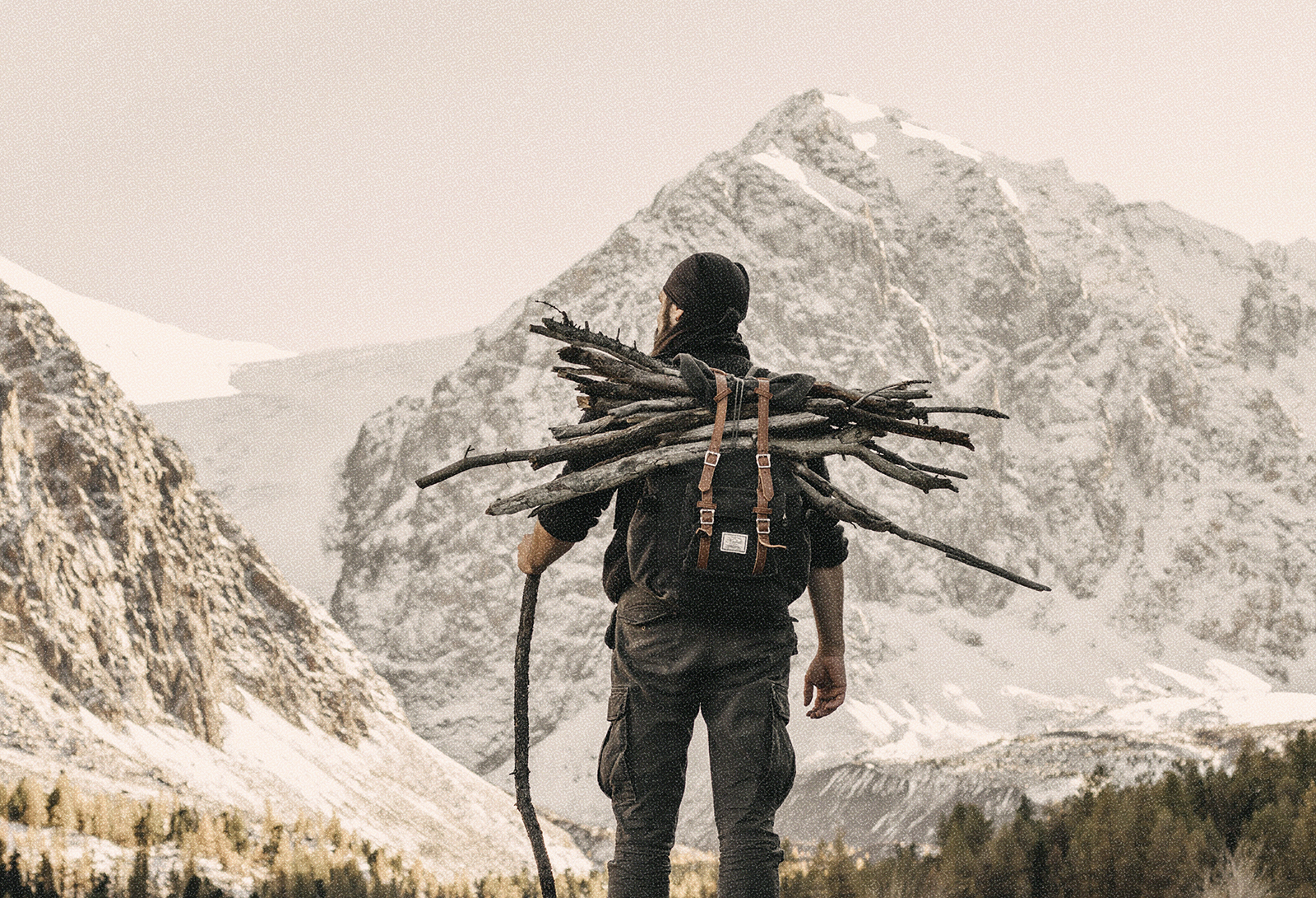 Image of a man in a beanie backpacking in snowy mountains with firewood stacked on his back and a walking stick in his hand