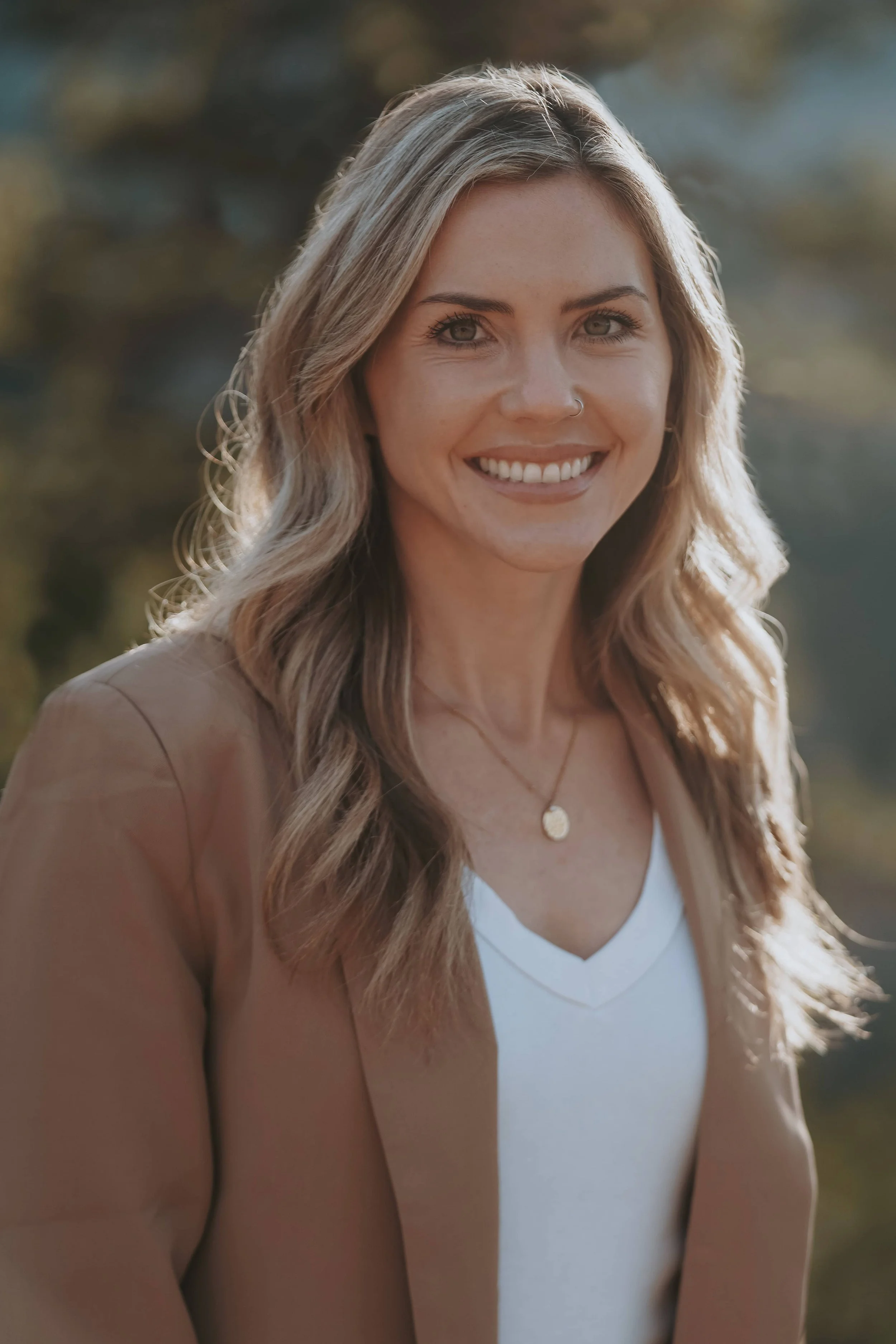 Skilled therapist smiling in nature looking at camera in white top with tan suit jacket.