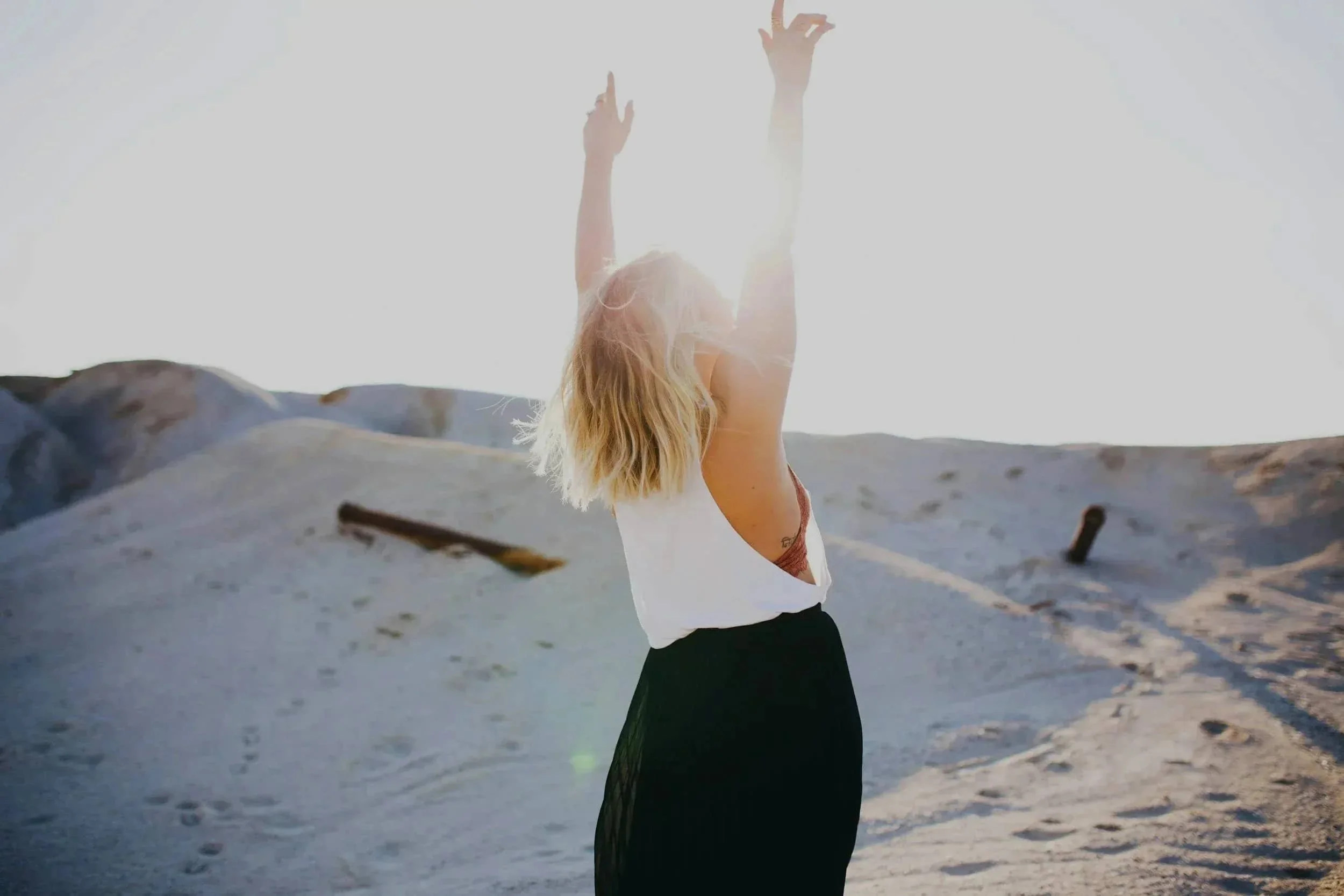 Woman with blonde hair wearing white top and black skirt raising her arms in a sandy outdoor area during sunset.