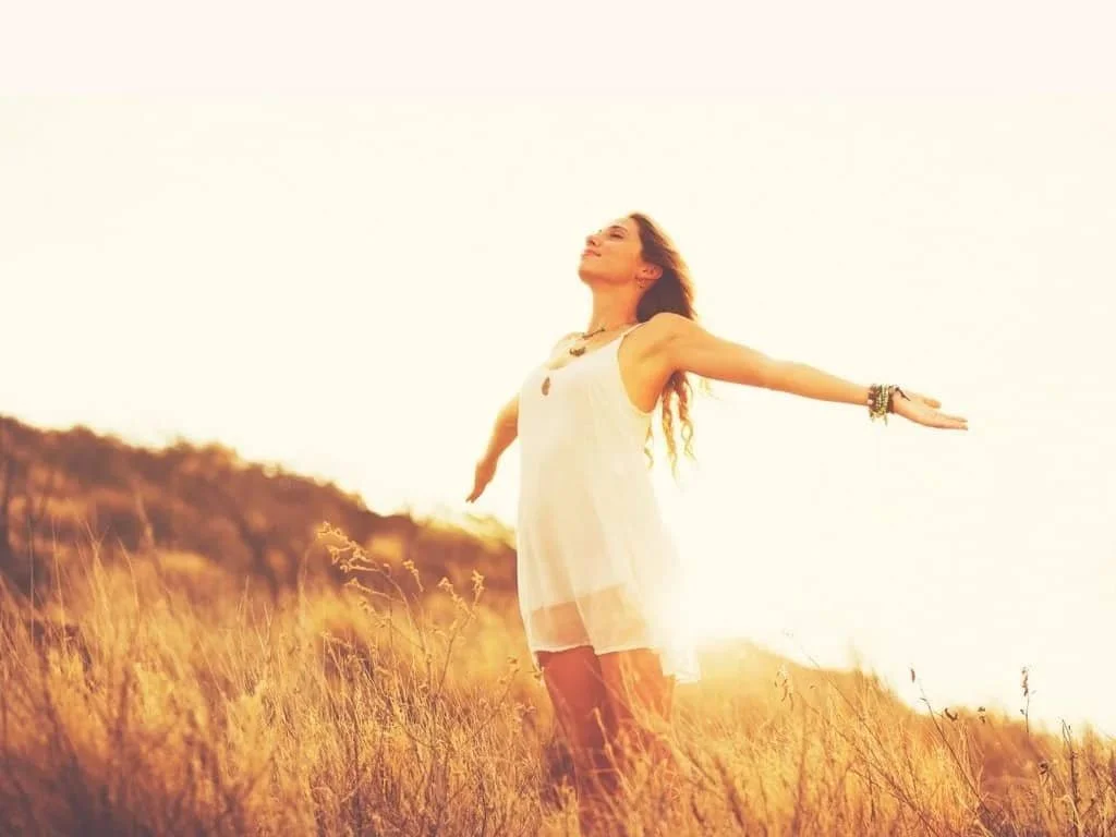 A woman stands outdoors in a field of tall grass with her arms outstretched and eyes closed. This woman has healed from trauma and anxiety after counseling in Denver Colorado.