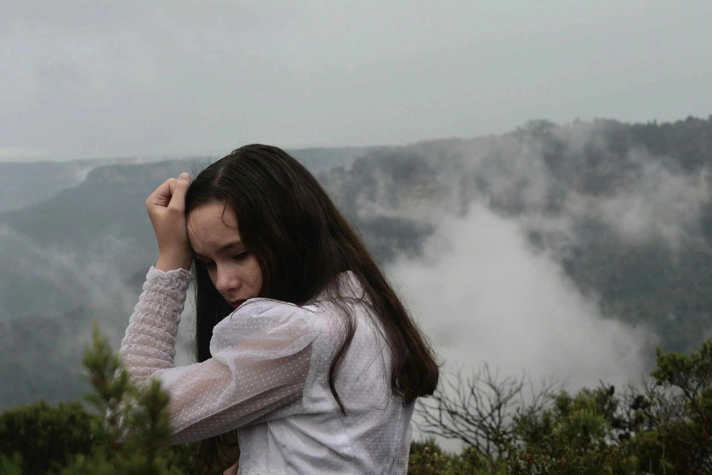 A young woman with long dark hair, wearing a white long-sleeve top, stands with her head tilted down and eyes closed, touching her head with one hand, in front of a foggy mountainous landscape. She has healed from her anxiety with therapy.