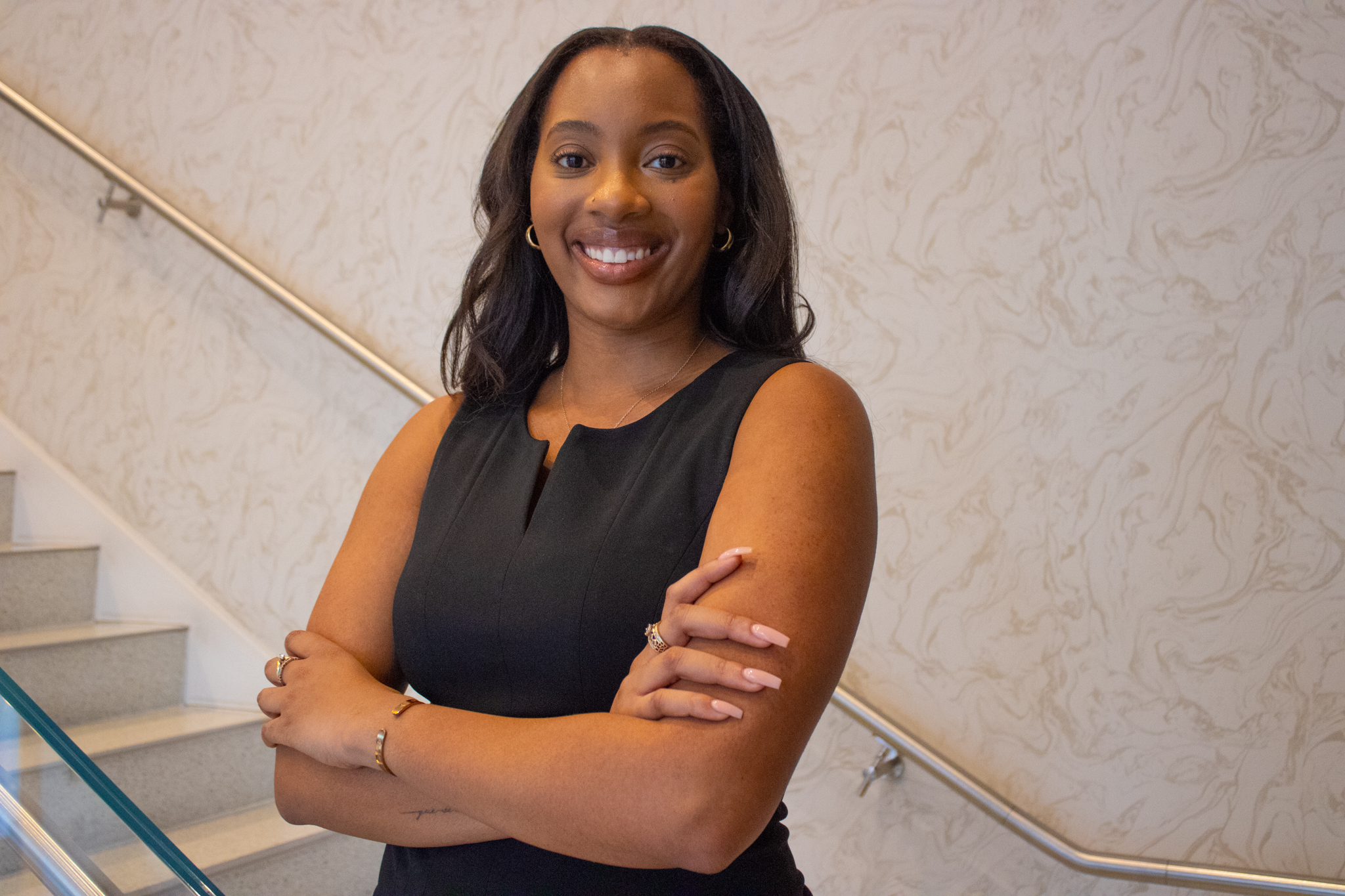 Jonea M. Weekley wearing a black sleeveless dress, standing with arms crossed in front of a patterned beige wall and staircase railing, smiling at the camera.