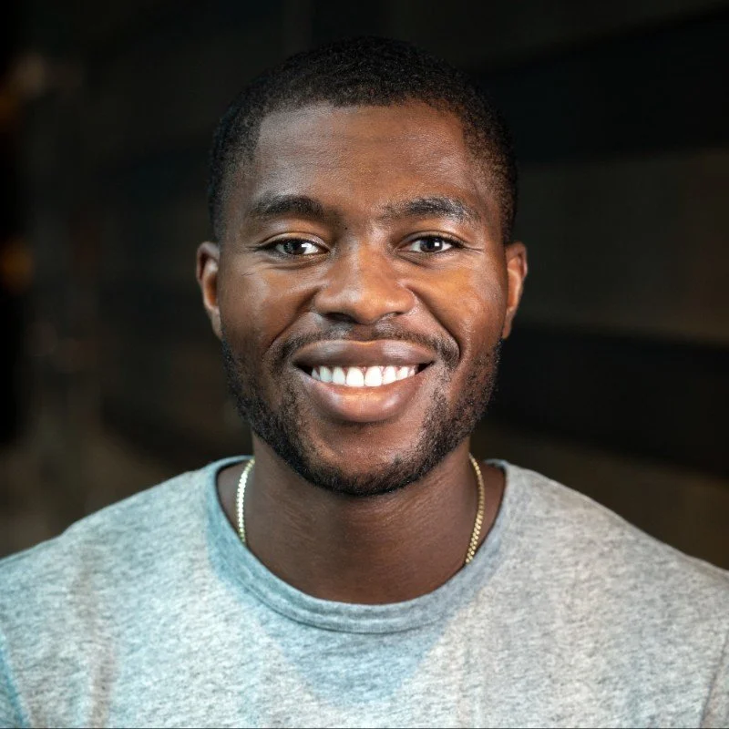 Close-up of a young smiling Black man with short hair, beard, wearing a gray t-shirt and a thin chain necklace.