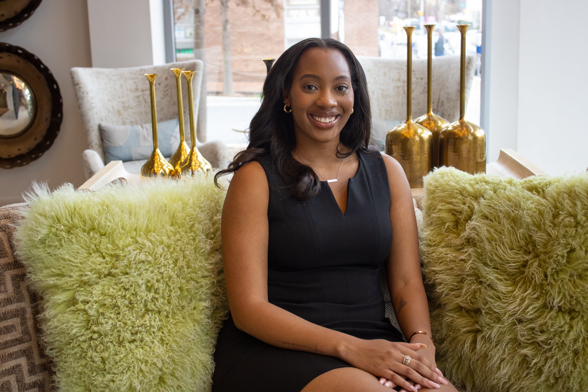 Jonea M. Weekley sitting on a patterned sofa with yellow furry pillows, smiling at the camera, wearing a black sleeveless dress, with gold decorative vases and large window in the background.