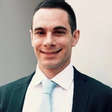A young man with dark hair, wearing a dark suit, white shirt, and light-colored tie, smiling at the camera against a plain light background.