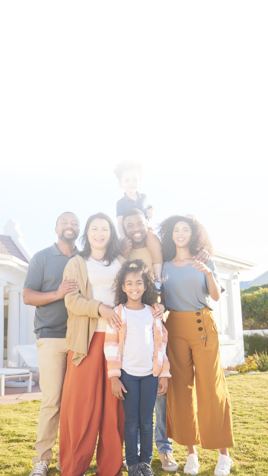A diverse family outdoor in front of white houses, smiling and enjoying the sunshine, including two adults, two children, and a young adult child on a man's shoulders.