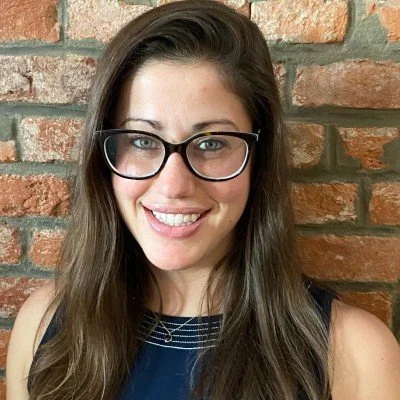 A woman with long brown hair wearing glasses and a dark top, smiling in front of a brick wall.