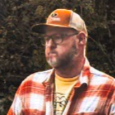 Man with glasses wearing orange and white trucker hat, plaid shirt, and yellow T-shirt outdoors. It is musician Jeff Thomas