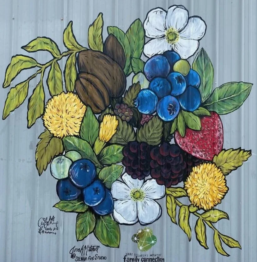 Vibrant mural of mixed fruits and flowers, featuring blueberries, strawberries, pecans, blackberries, yellow flowers, and white blossoms with green leaves, painted on a wall.