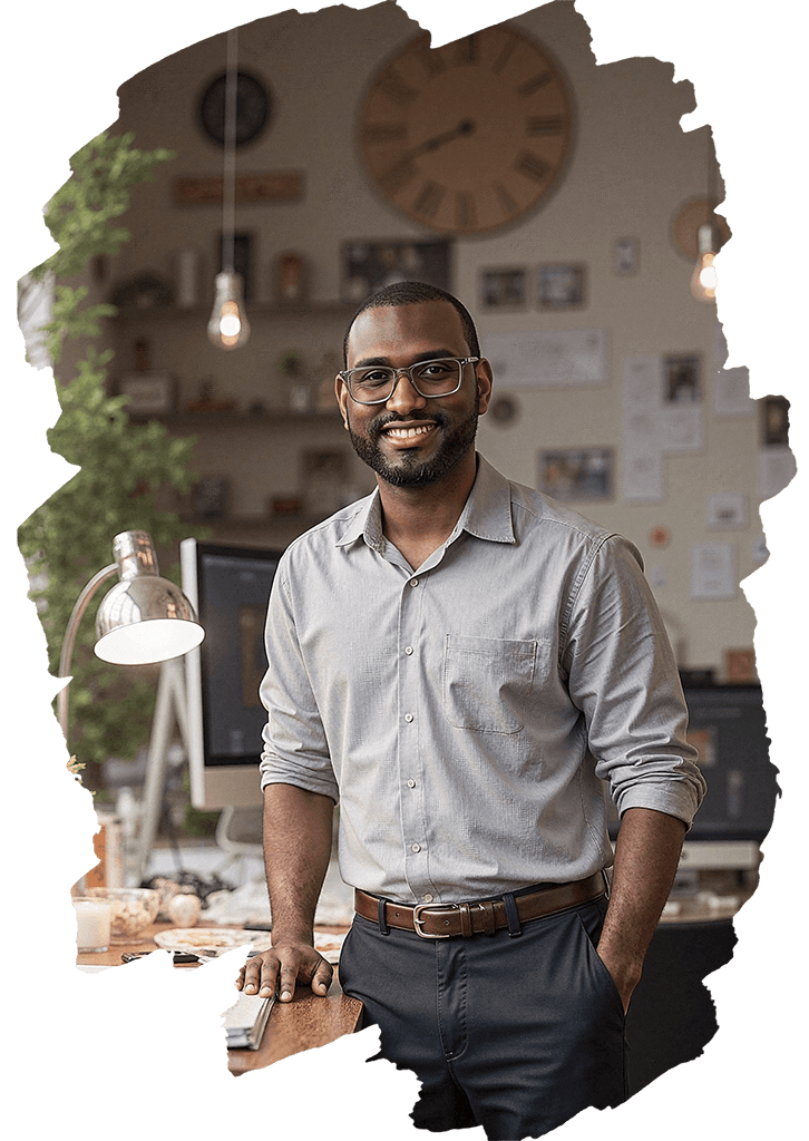 A man wearing glasses and a light gray shirt, smiling, standing in an office environment with a large wall clock, a computer, and decorative items in the background.