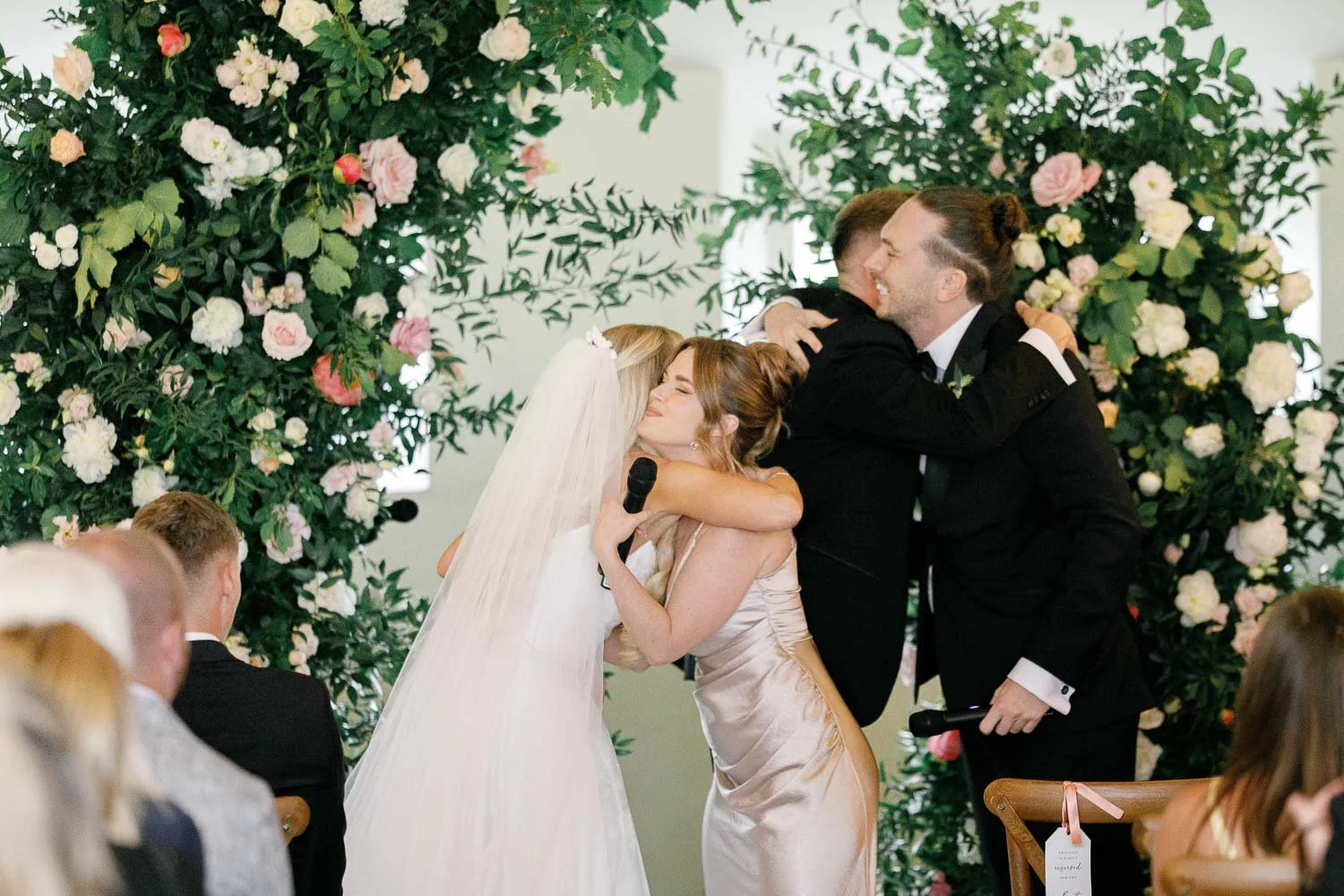 bride and groom simultaniously hugging their best man and maid of honour in front of floral arch