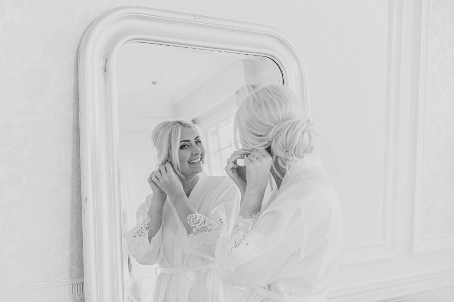 Black and white photo of bride putting on earrings in mirror