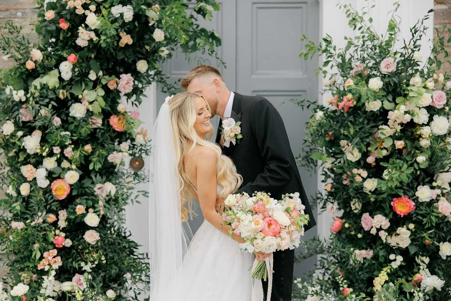 Bride and groom surrounded by big floral arch in front of the entrance to Boconnoc Estate