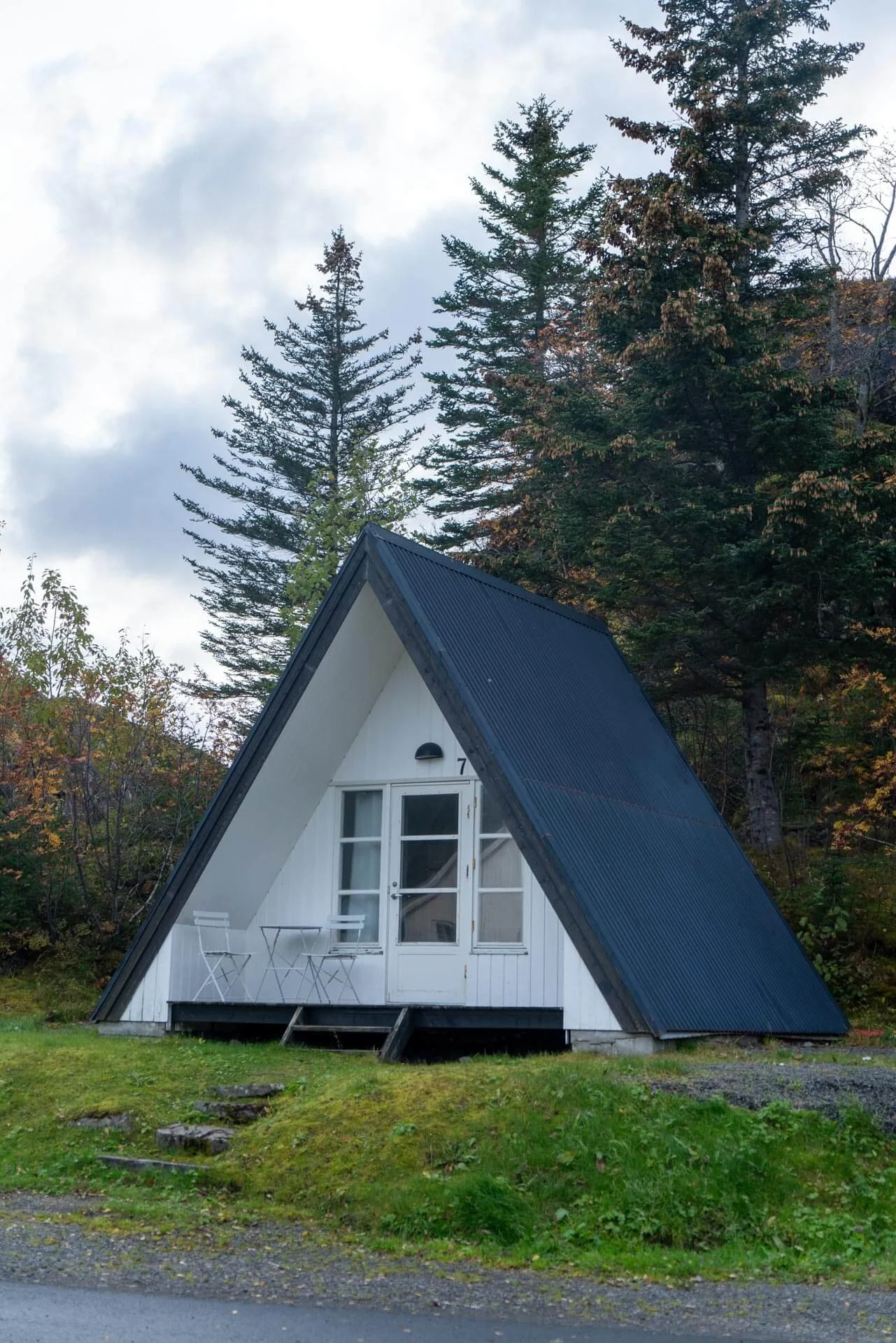 A small A-frame house with white walls and a black metal roof, surrounded by trees and greenery.