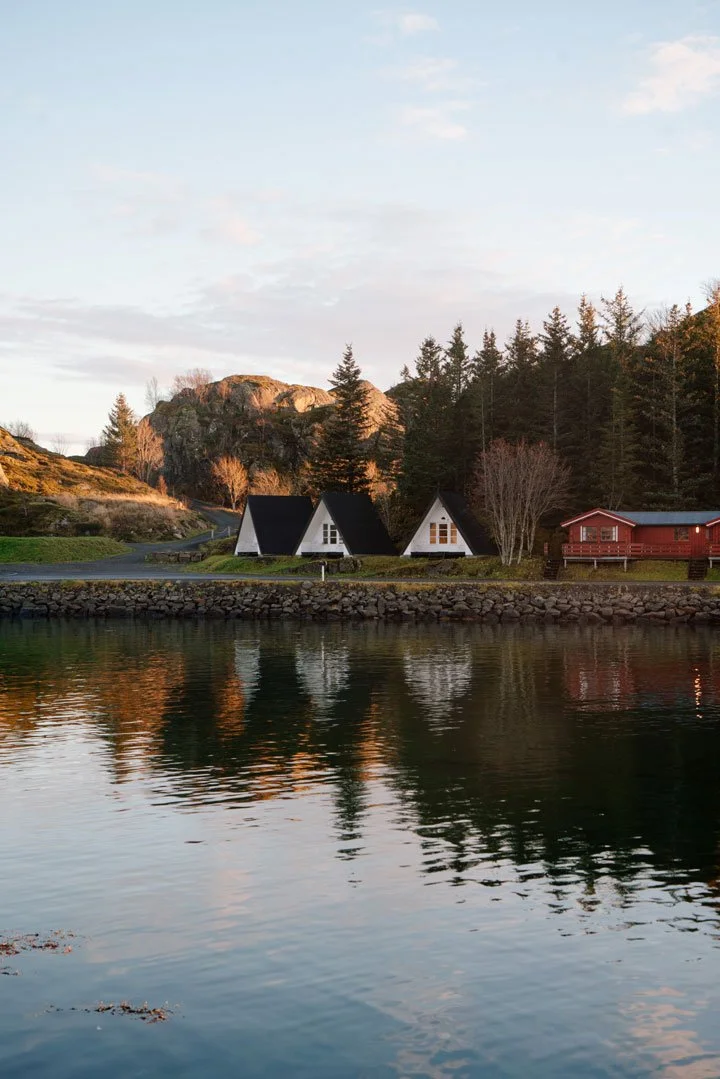 Scenic view of a lakeside with three small white A-frame houses, a larger red building, dense trees, and rocky hills in the background during sunset.