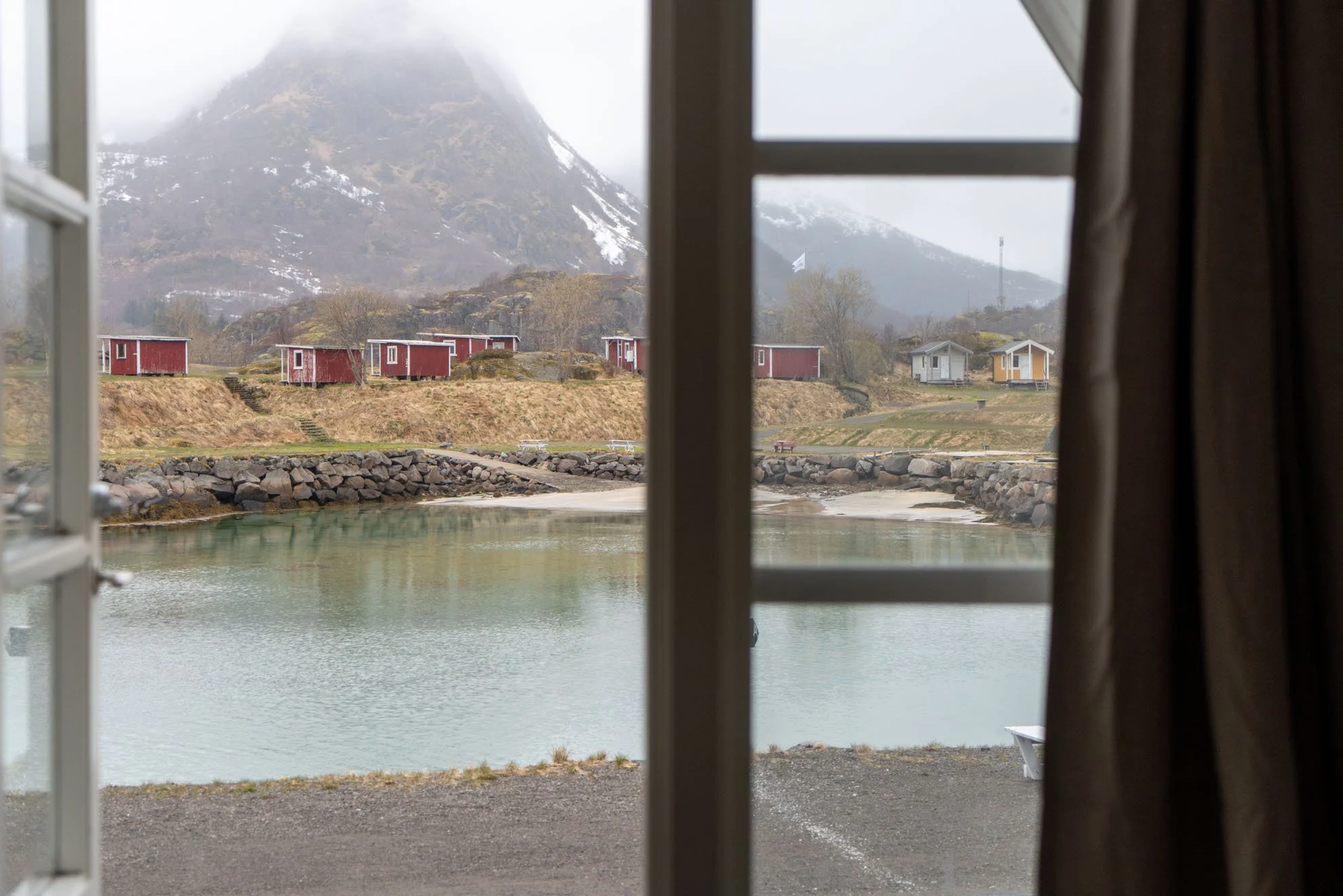 View of a lake with a rocky shoreline, small houses on a grassy hill, and snow-capped mountains in the background, seen through a window with curtains.
