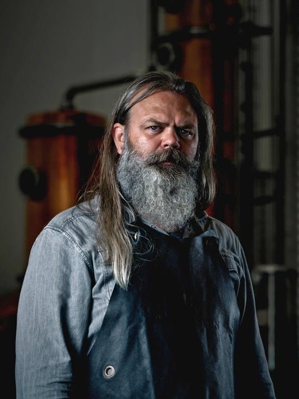 A man with long gray hair and a gray beard, wearing a denim shirt and black apron, standing in a workshop with tools in the background.