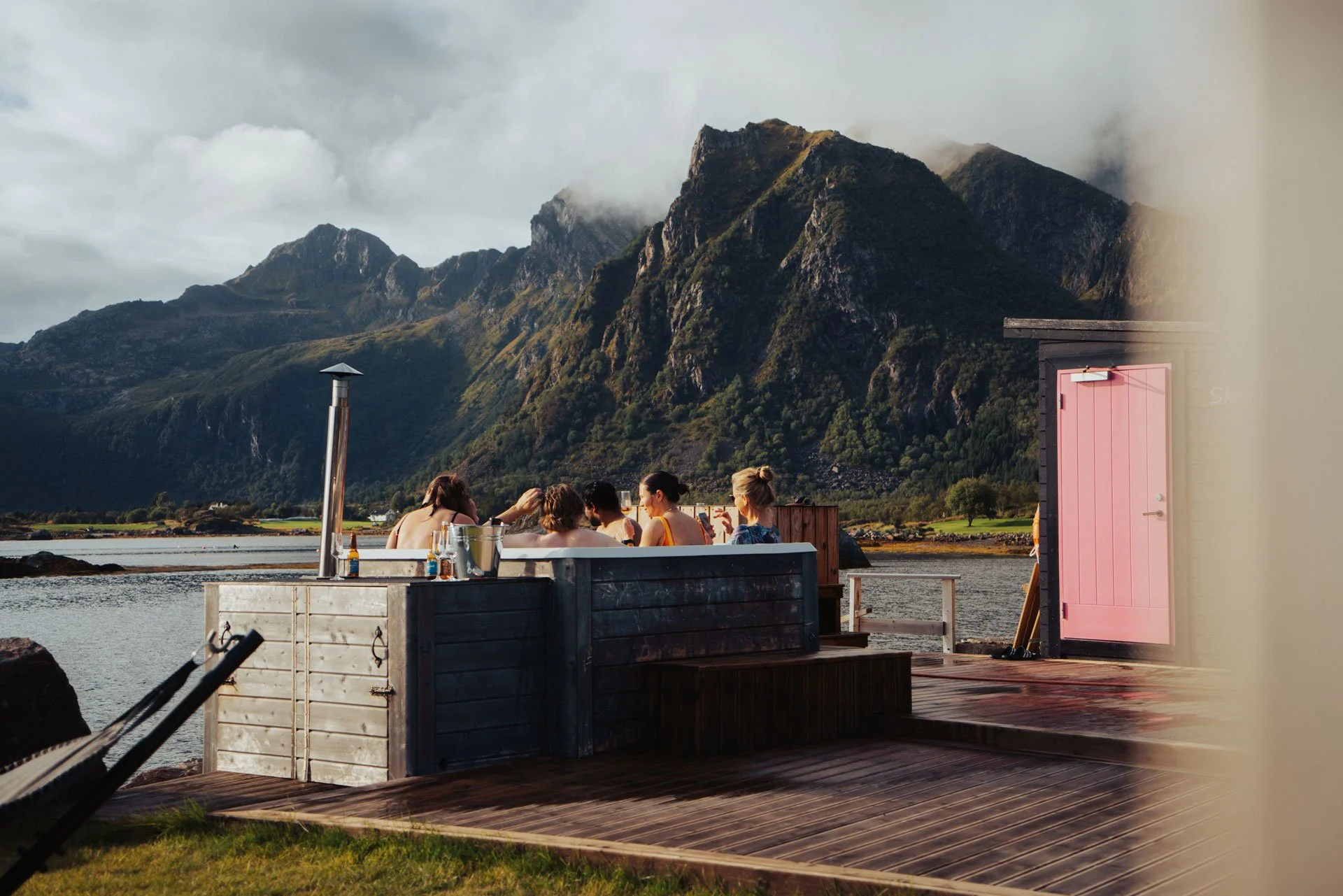 People relaxing in an outdoor hot tub beside a lake with mountains in the background