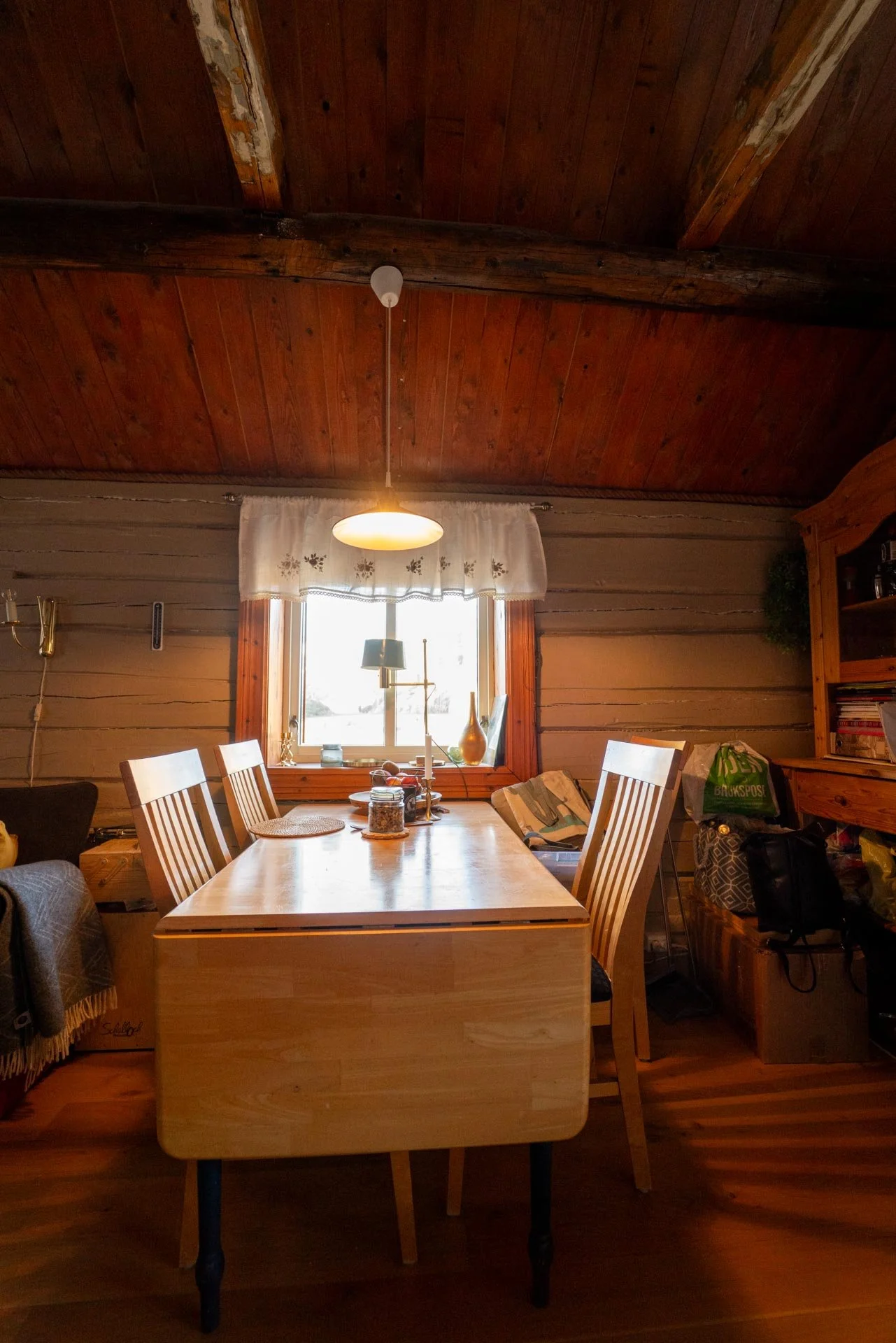 A cozy dining room with a wooden ceiling and wall, a window with white curtains, a wooden table with chairs, and various decorative items and clutter on and around the table.