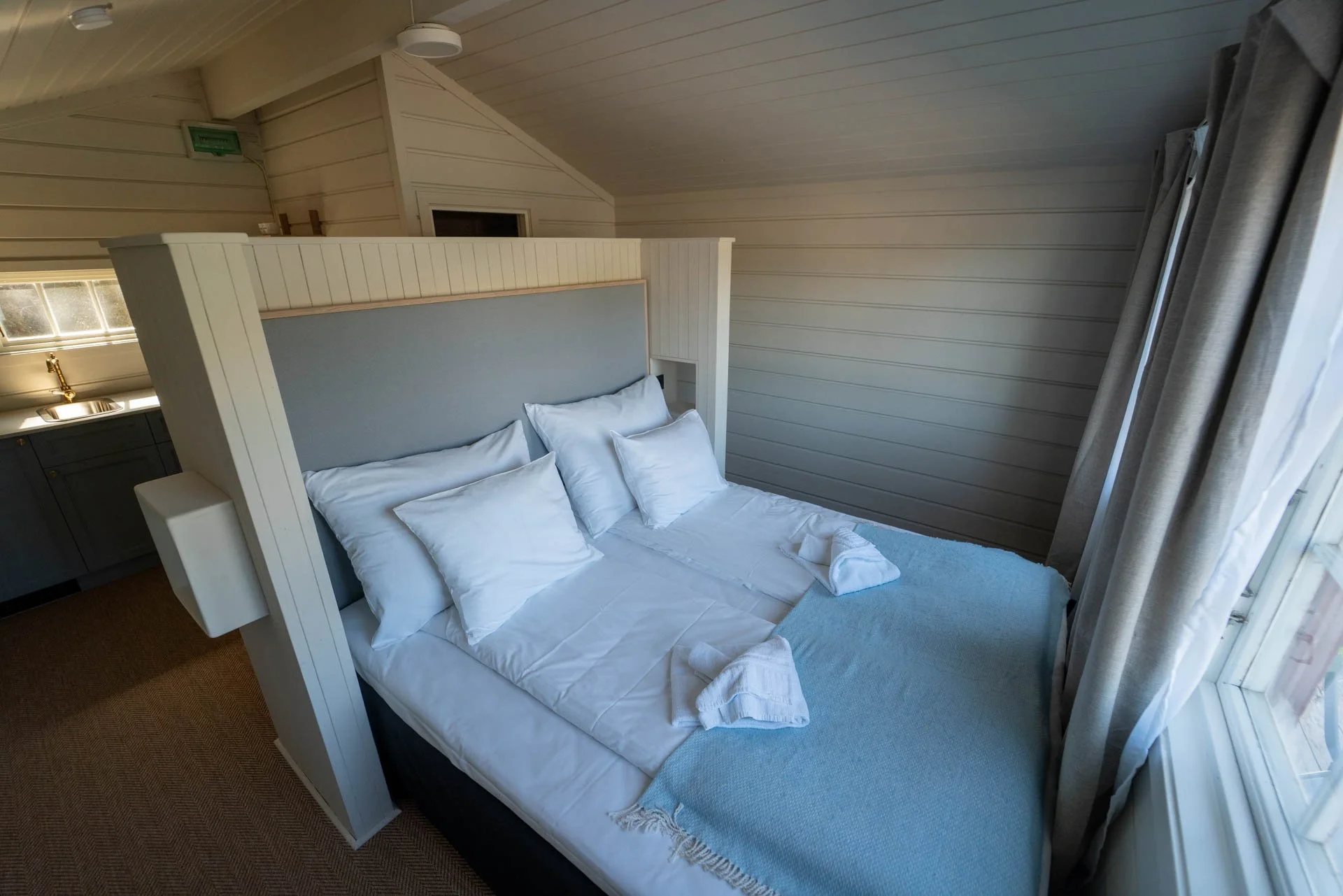 A neatly made bed with white sheets and multiple pillows in a cozy room with wooden panel walls and a ceiling, next to a window with curtains.