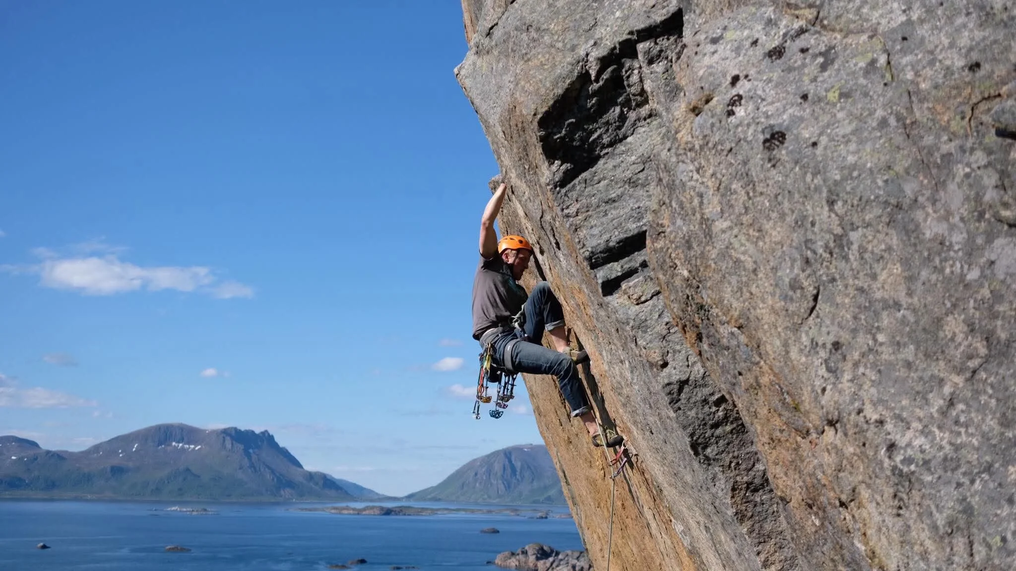 Person wearing an orange helmet rock climbing on a steep outdoor cliffside with a body of water and distant mountains in the background.