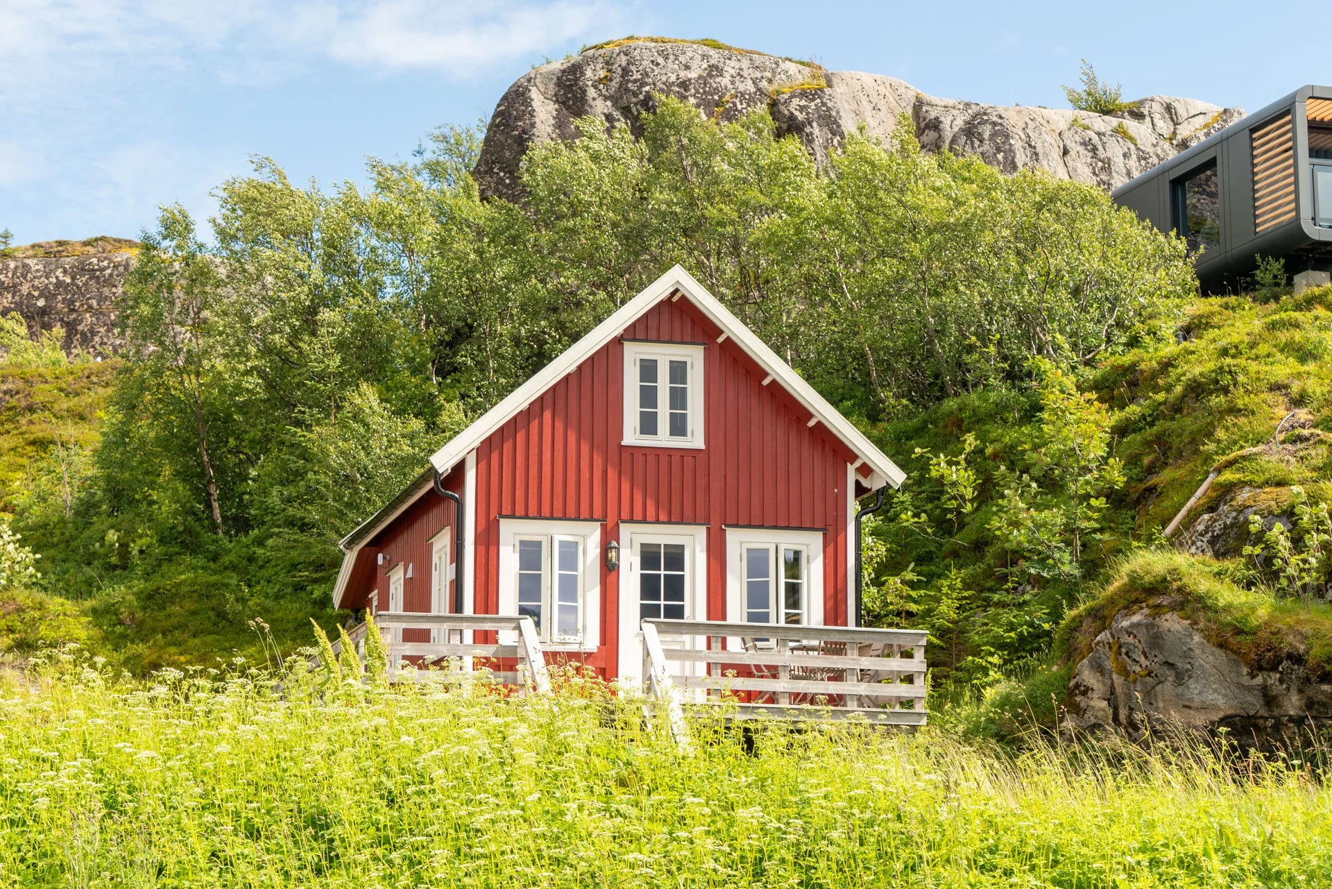 Red wooden house with white trim, a deck with outdoor furniture, on a hillside with trees and large rocks in the background.