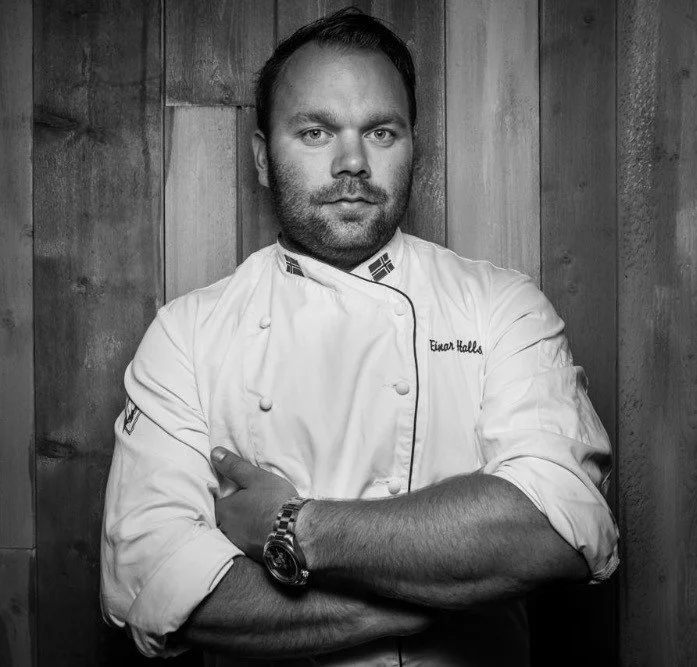 Black and white photo of a male chef in a white chef's jacket standing in front of a wooden wall, arms crossed, looking at the camera.