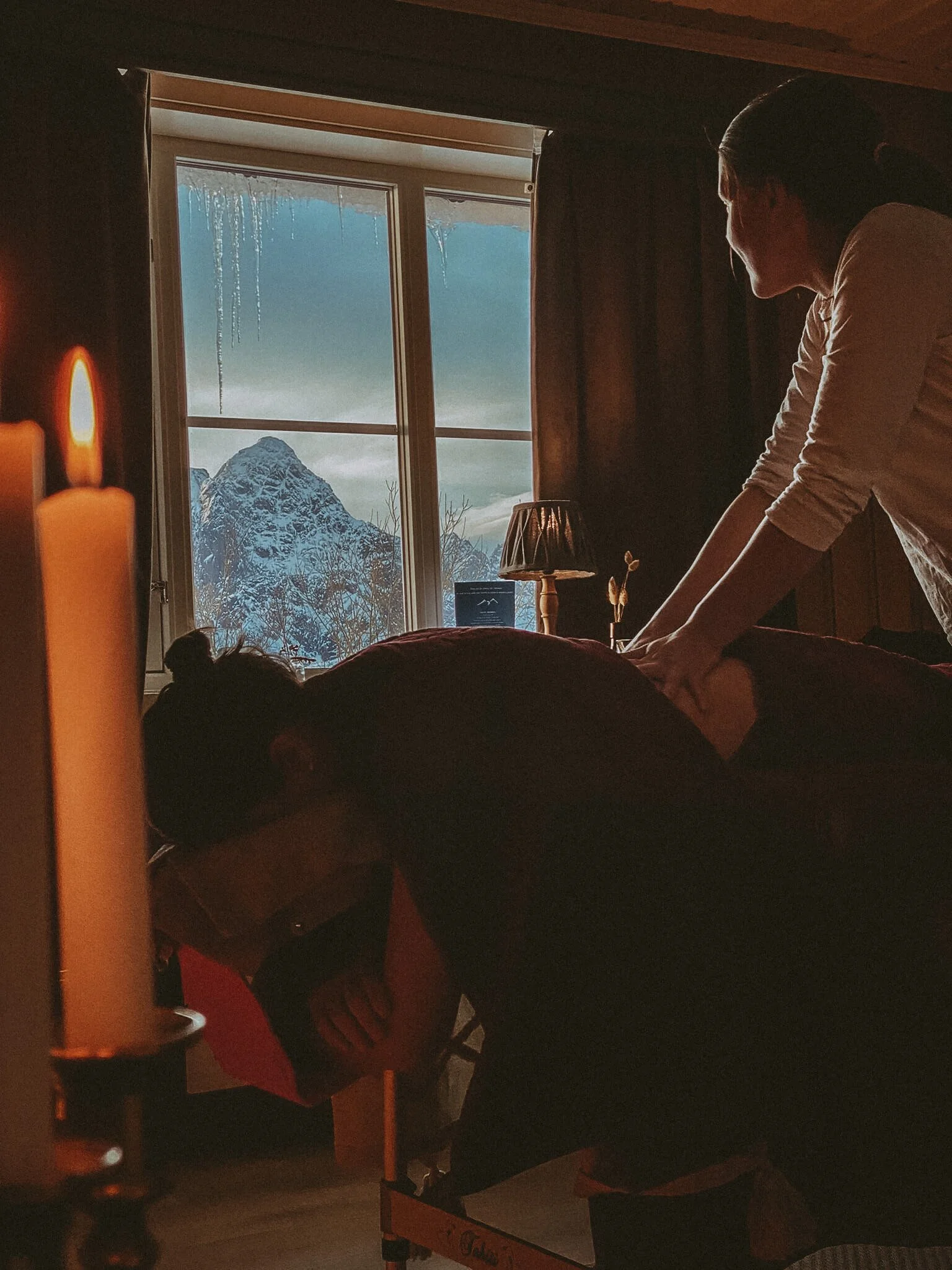 A woman giving a massage to a person lying face down on a massage table inside a cozy, dimly lit room with a window showing a snowy mountain landscape outside.