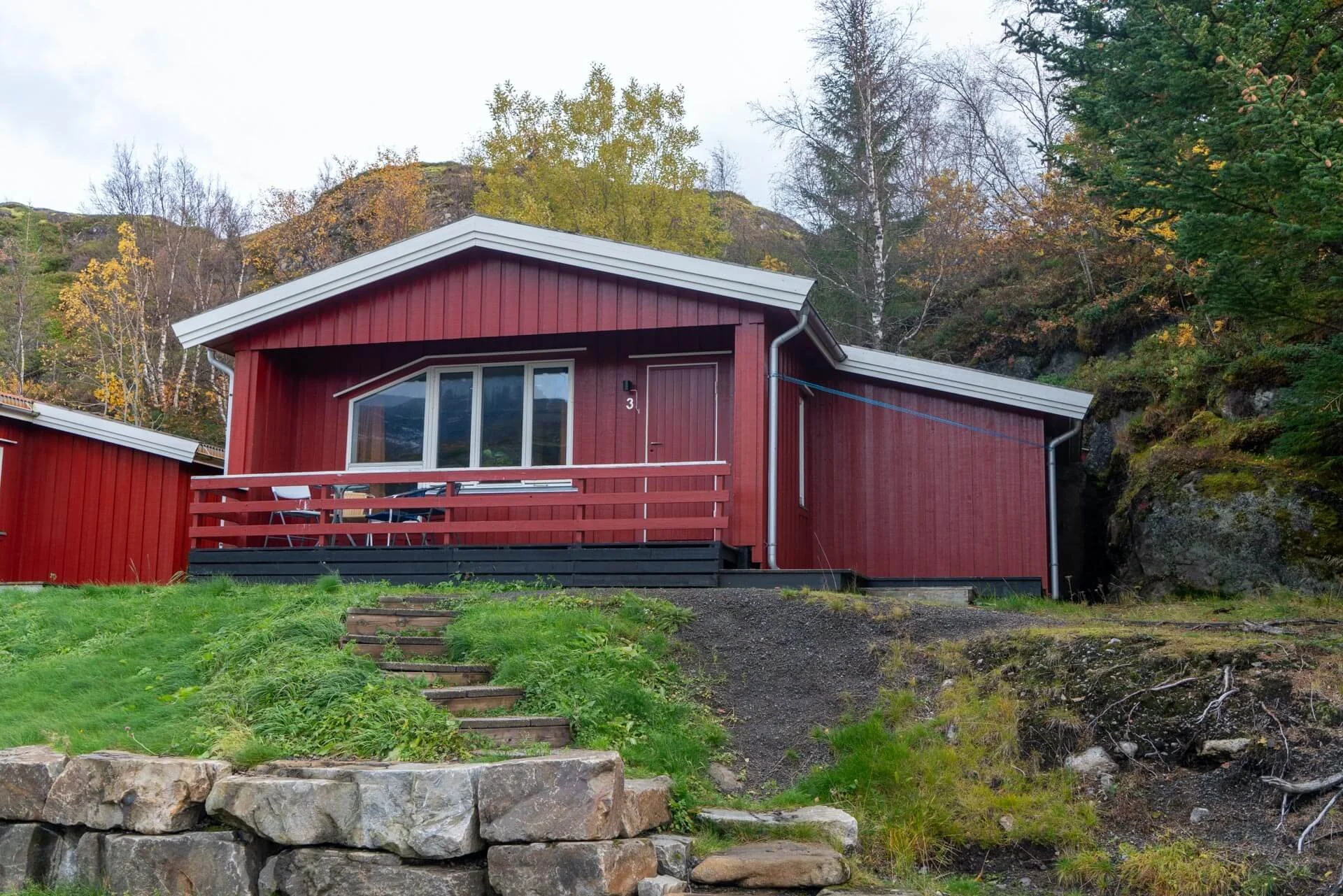 Red house with a small deck and steps leading up a grassy hill, surrounded by trees with autumn foliage and rock formations in the background.