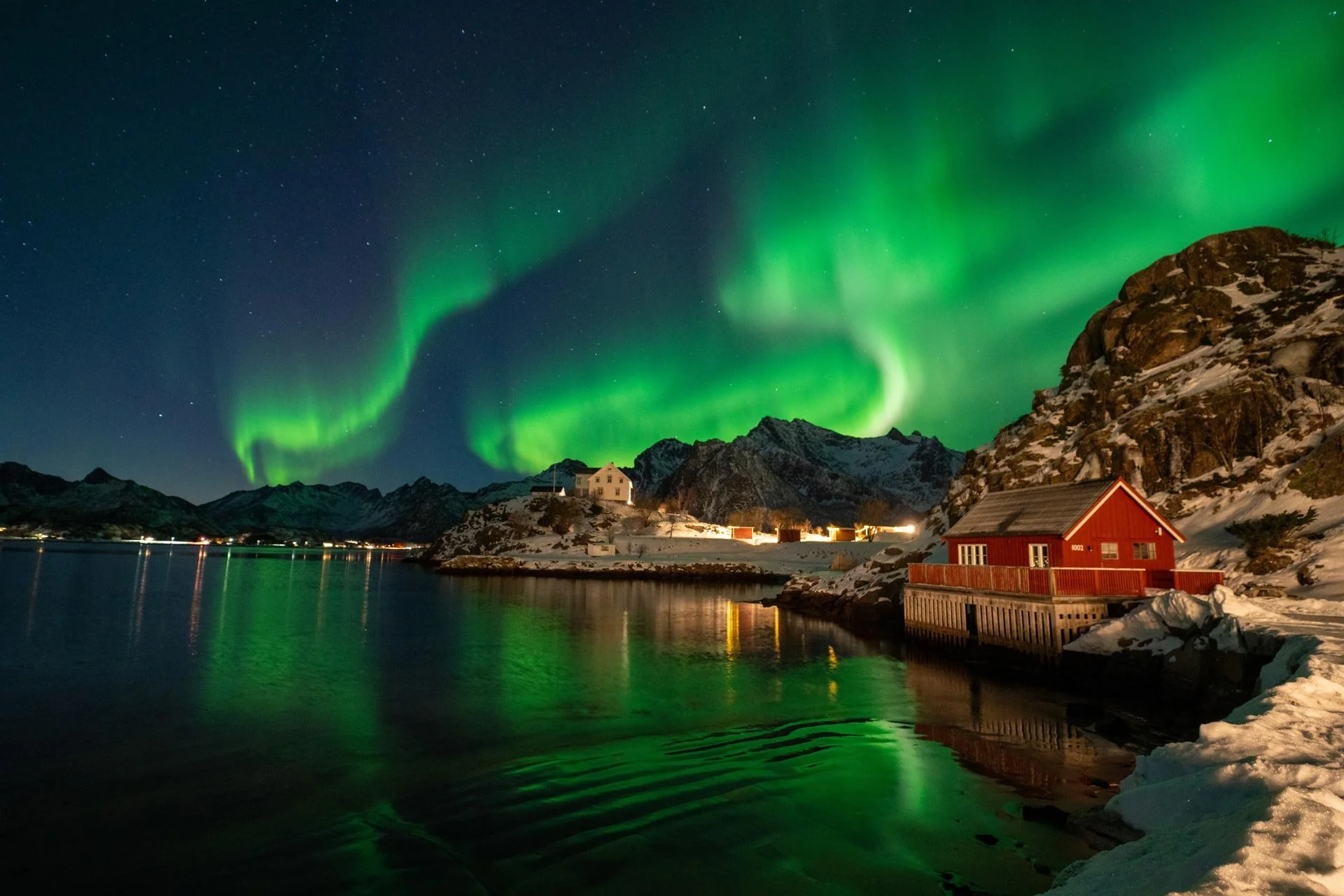 A red wooden house on stilts by a rocky shoreline with a small sandy beach, surrounded by mountainous terrain under a cloudy sky.