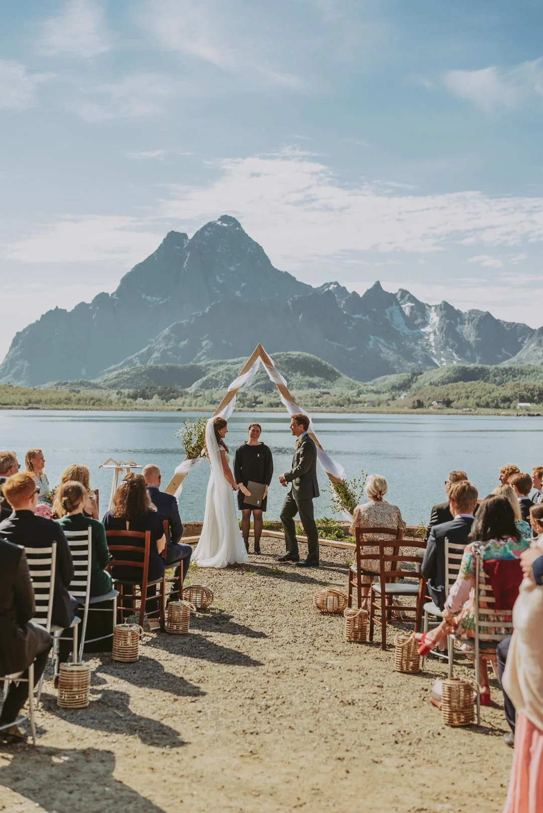 A wedding ceremony taking place outdoors by a lake with tall mountain peaks in the background, featuring the bride and groom exchanging vows under a triangular wooden arch decorated with white fabric and greenery, with guests seated on either side.