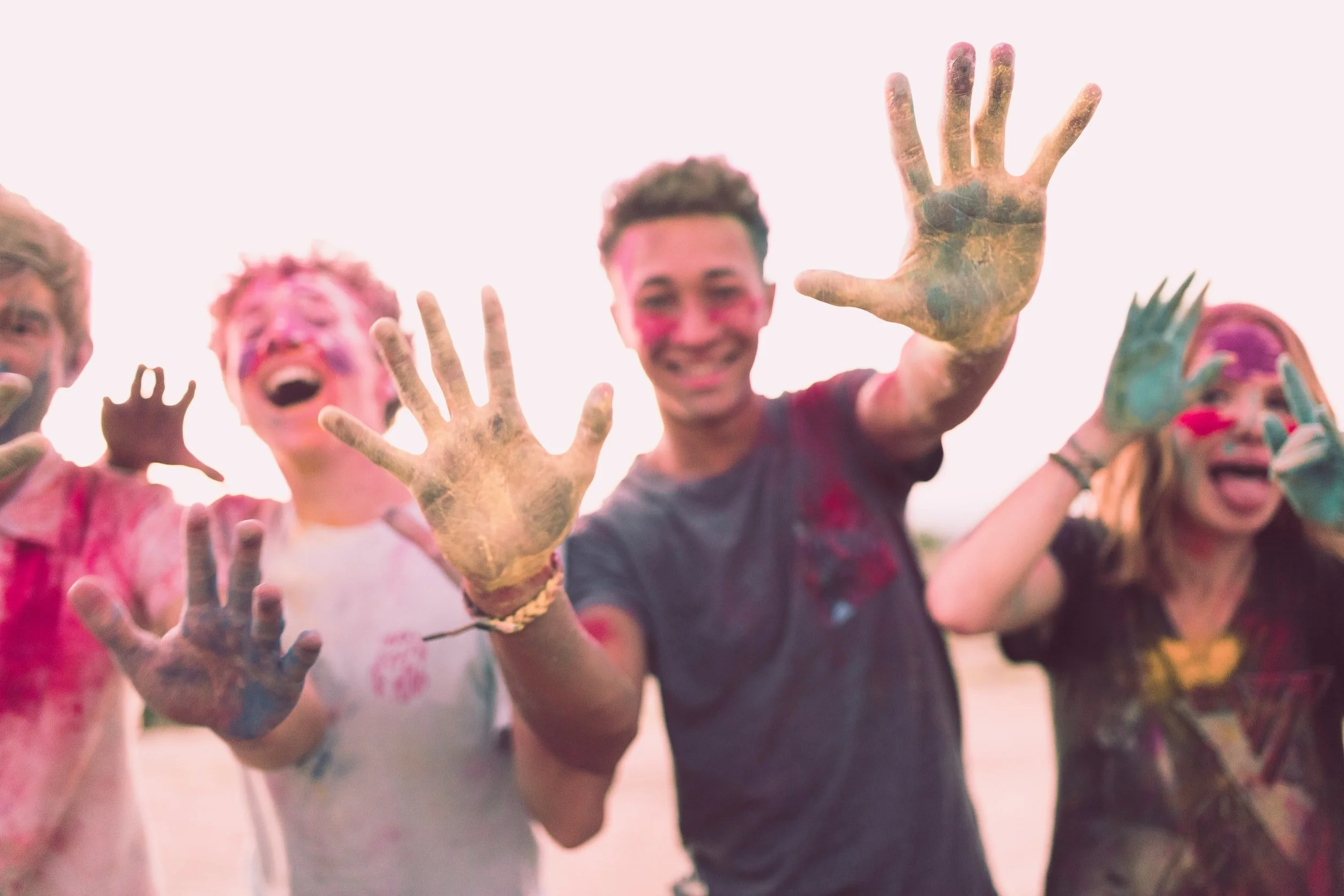 Aesthetic image of 3 white cis male and 1 white cis female (all aged in early 20's) covered in paint holding up hands, pulling faces and generally having fun