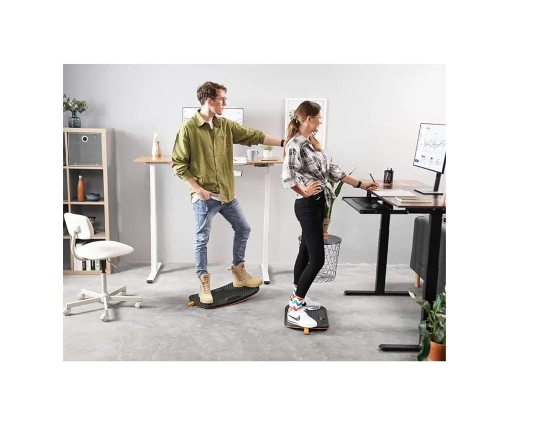 Two young adults, a man and a woman, using balance boards on a treadmill and a standing desk in an office, with the man holding the woman's shoulder for support.