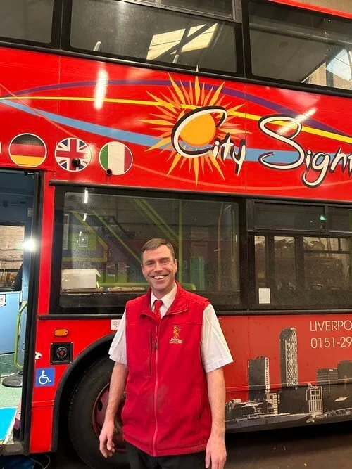 A man in a red uniform standing in front of a red double-decker city sightseeing bus with various flags and the word 'City Sightseeing' on it.