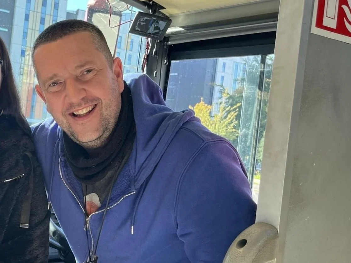 Smiling man wearing a blue hoodie inside a bus with city buildings visible through windows.