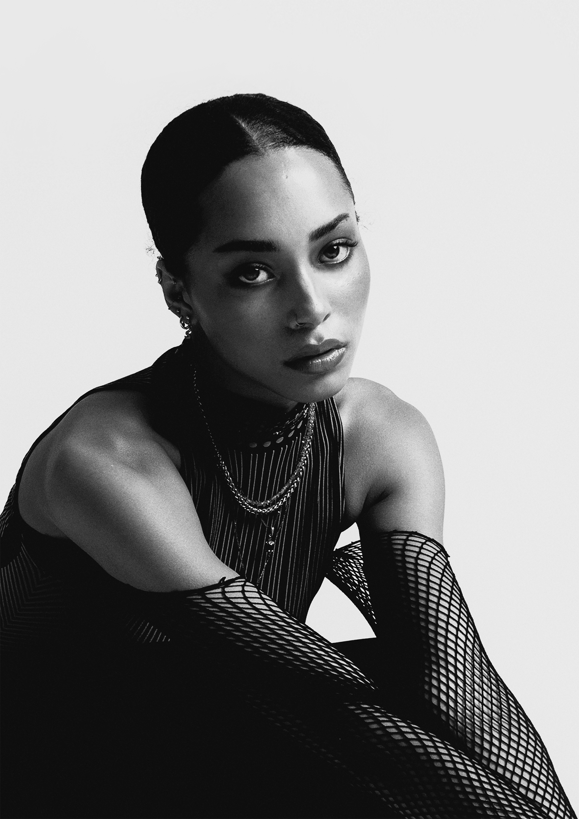 Black-and-white portrait of a woman with short hair, wearing fishnet stockings and layered necklaces, posed against a plain white background.