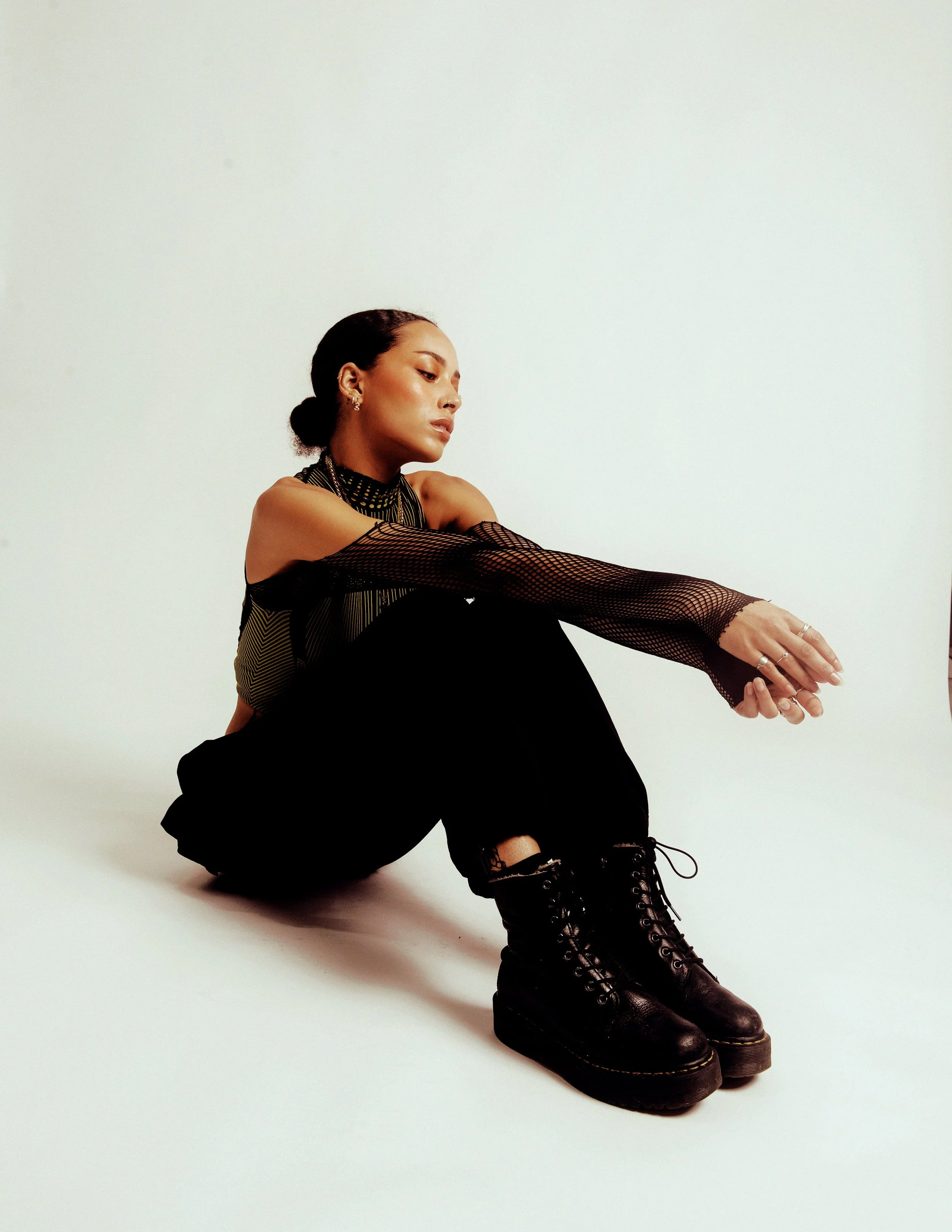 A woman with dark hair tied back, wearing a black mesh long-sleeve top and black pants, sitting on the floor with legs extended, wearing black lace-up boots, against a white background.