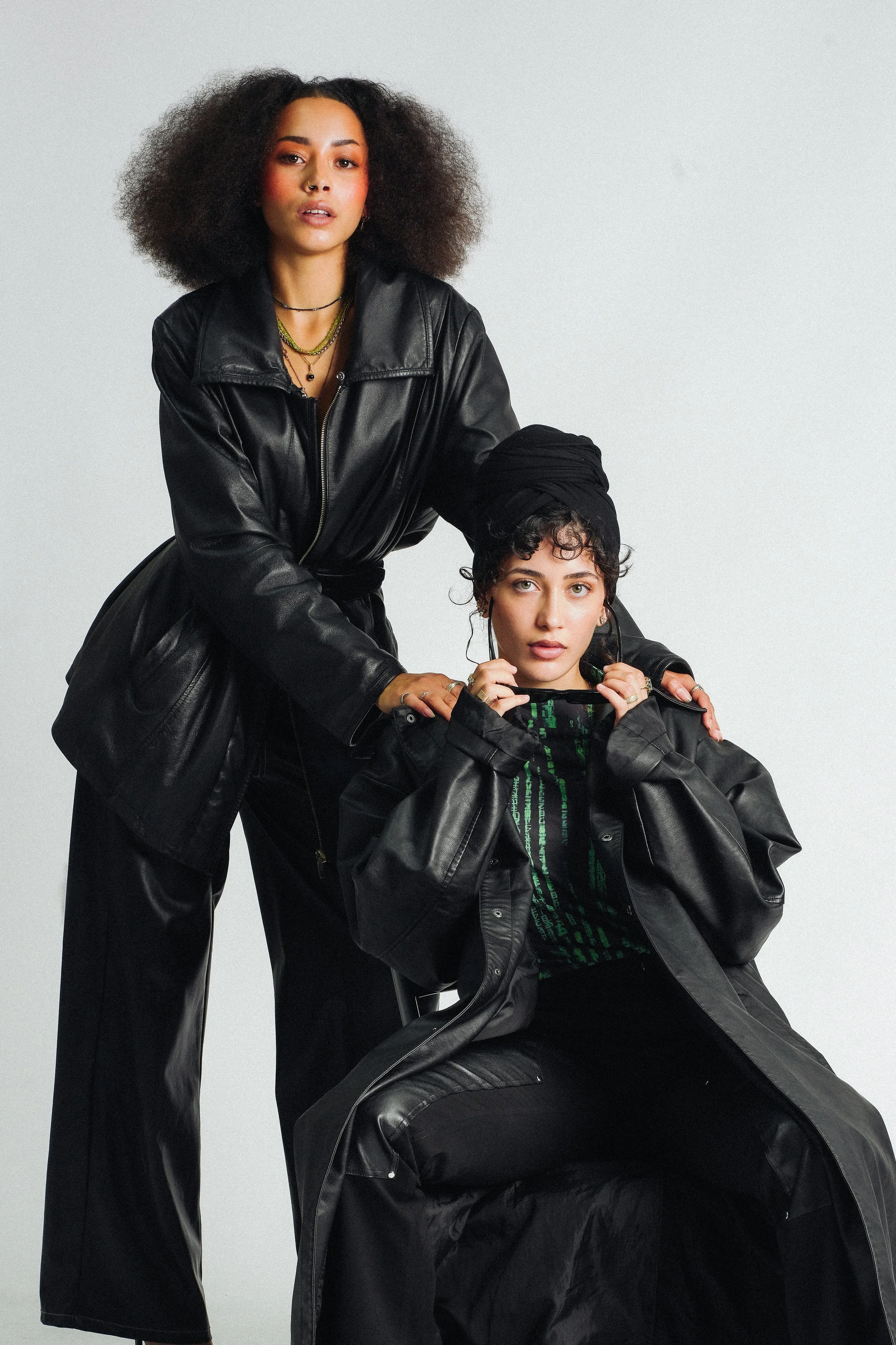 Two women posing together wearing black leather jackets, one standing behind and the other sitting, both with short curly hair and wearing layered necklaces.