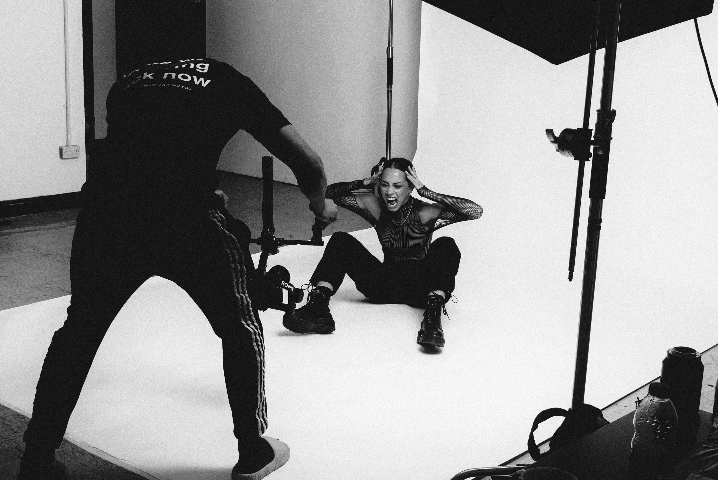 A photo shoot in a studio featuring a woman with styled hair and dark clothing sitting on the floor and screaming with her hands on her head, while a man in athletic wear is adjusting equipment nearby, with studio lights and a plain white backdrop in the background.