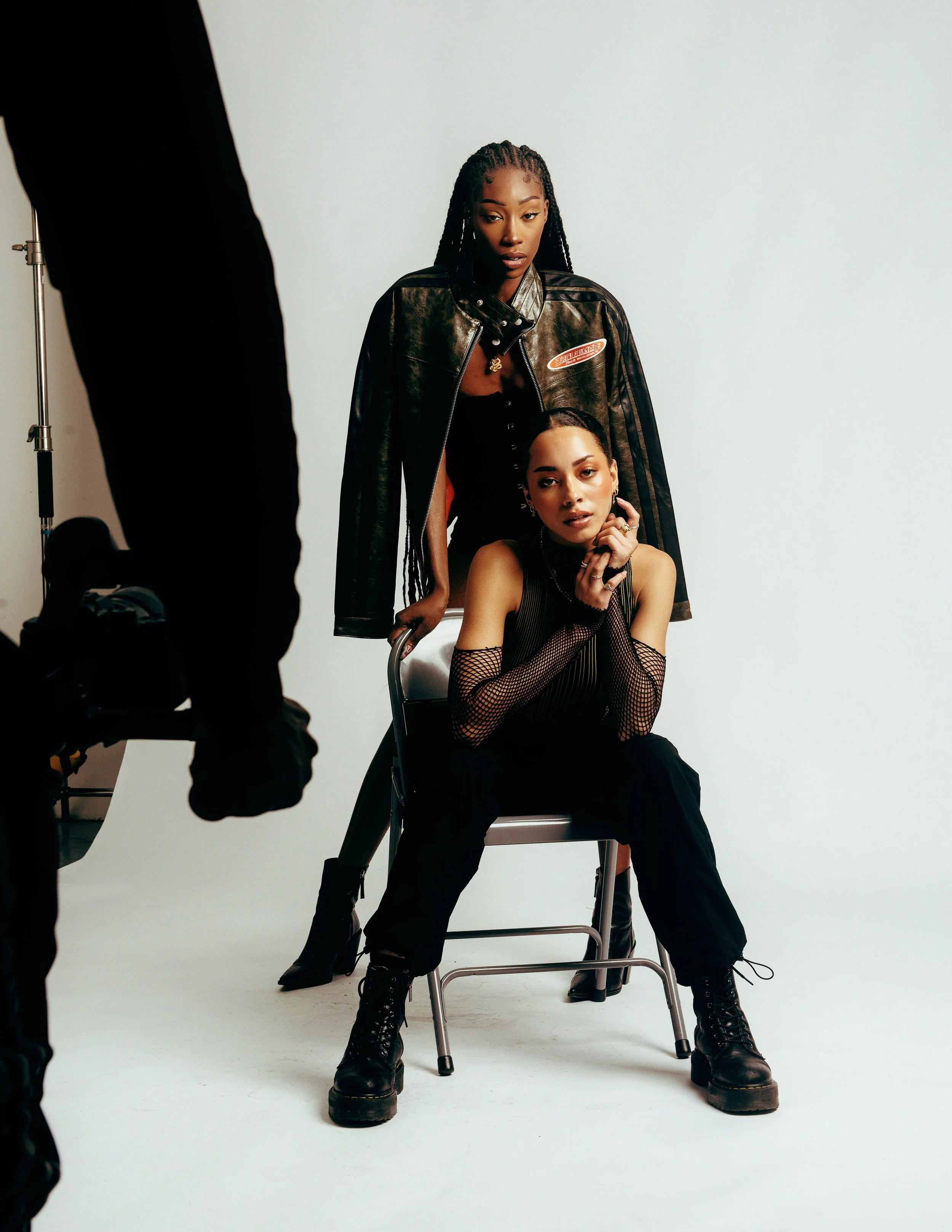 Two young women with dark hair and dramatic makeup posing in a photo studio. One sitting on a chair, wearing fishnet gloves and black boots, the other standing behind her, wearing a black jacket with patches and black pants. A person in black is visi