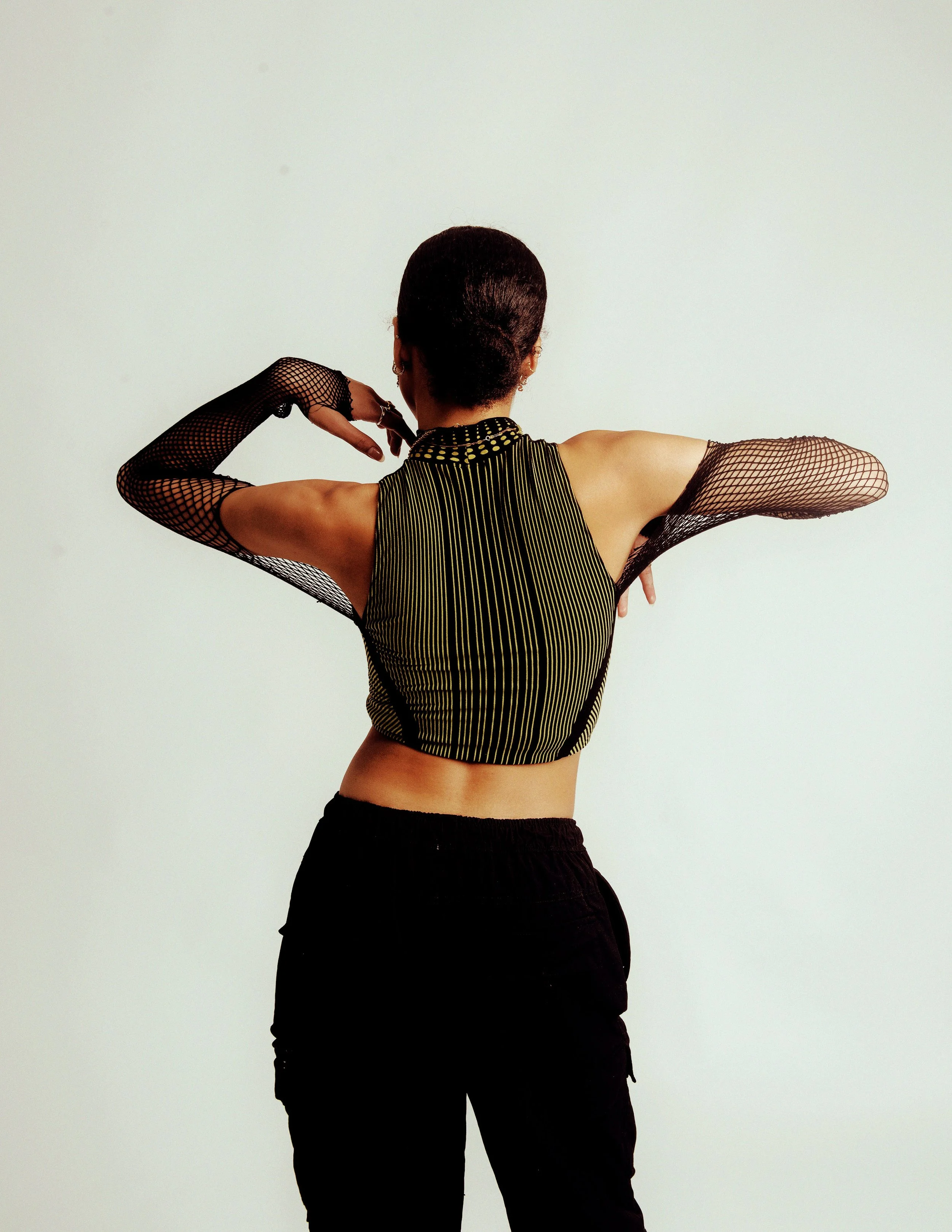 Back view of a woman in a sleeveless, black and yellow striped top with mesh sleeves, black shorts, and earrings, posing with arms raised and hands near her head against a plain white background.