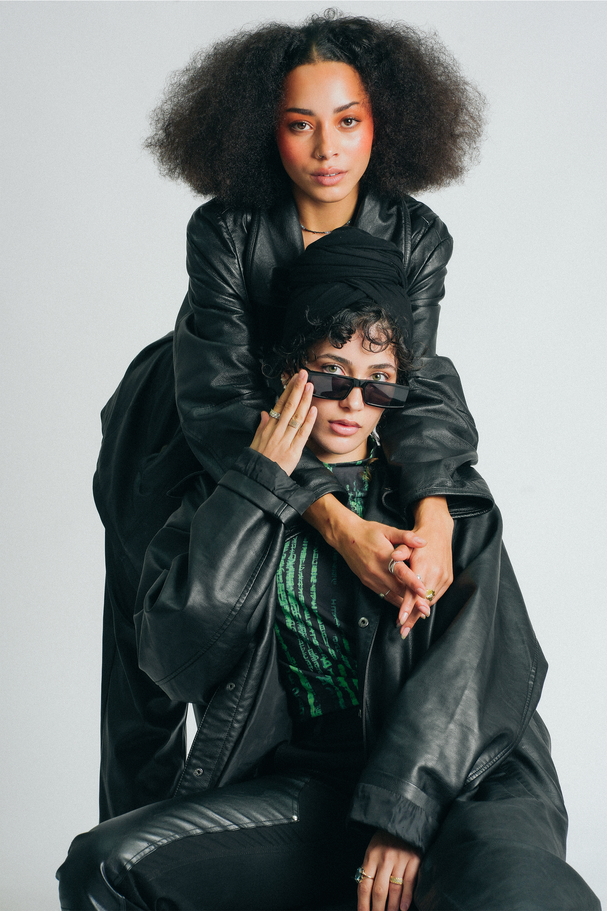 Two individuals dressed in black leather jackets posing together against a plain white background. The person on top is a woman with voluminous curly hair, light makeup, and a nose ring, looking directly at the camera. The person below, also with cur