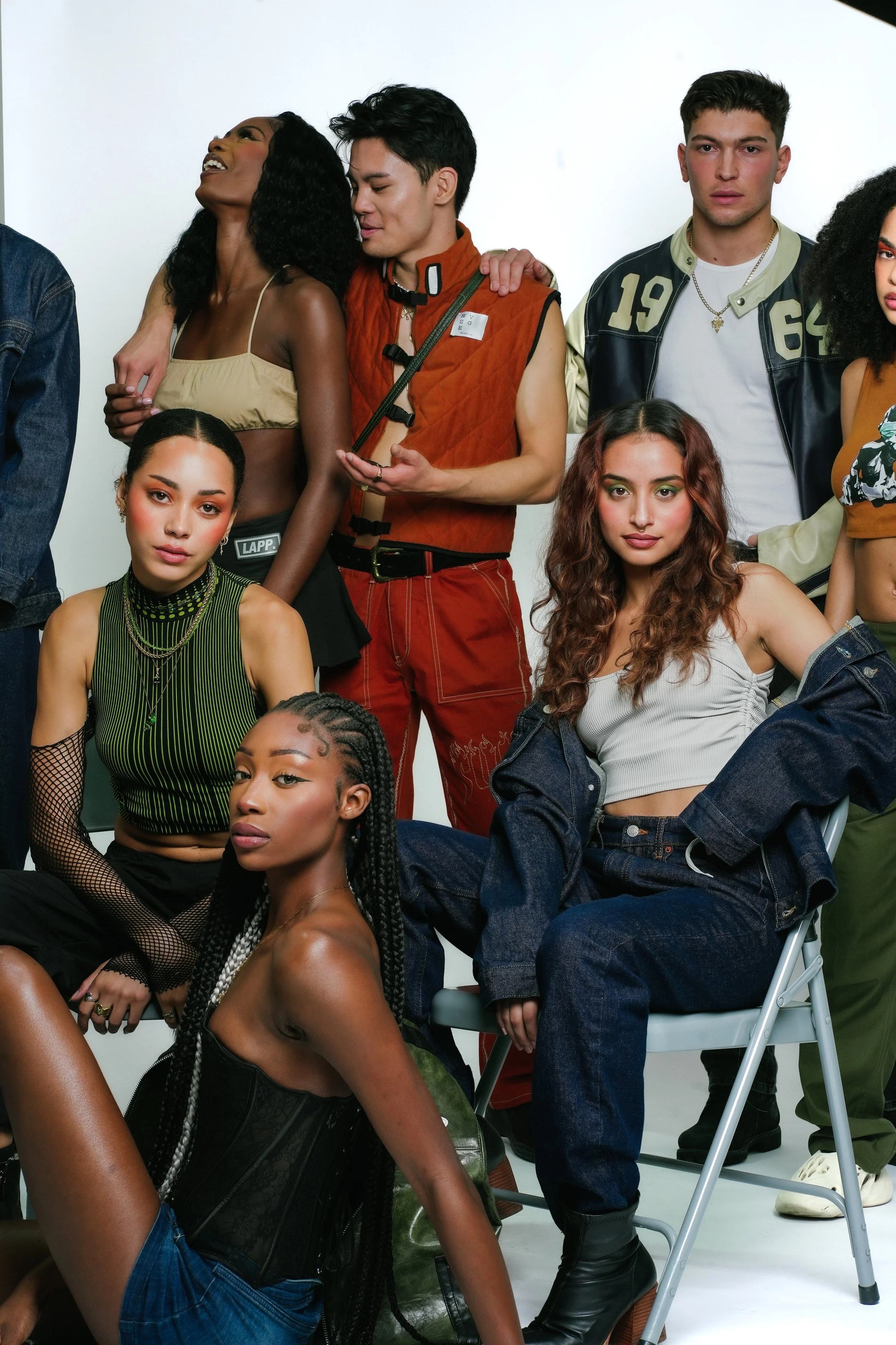 Group of diverse young adults posing for a photo against a white background, with some sitting and others standing, displaying various fashionable outfits and hairstyles.