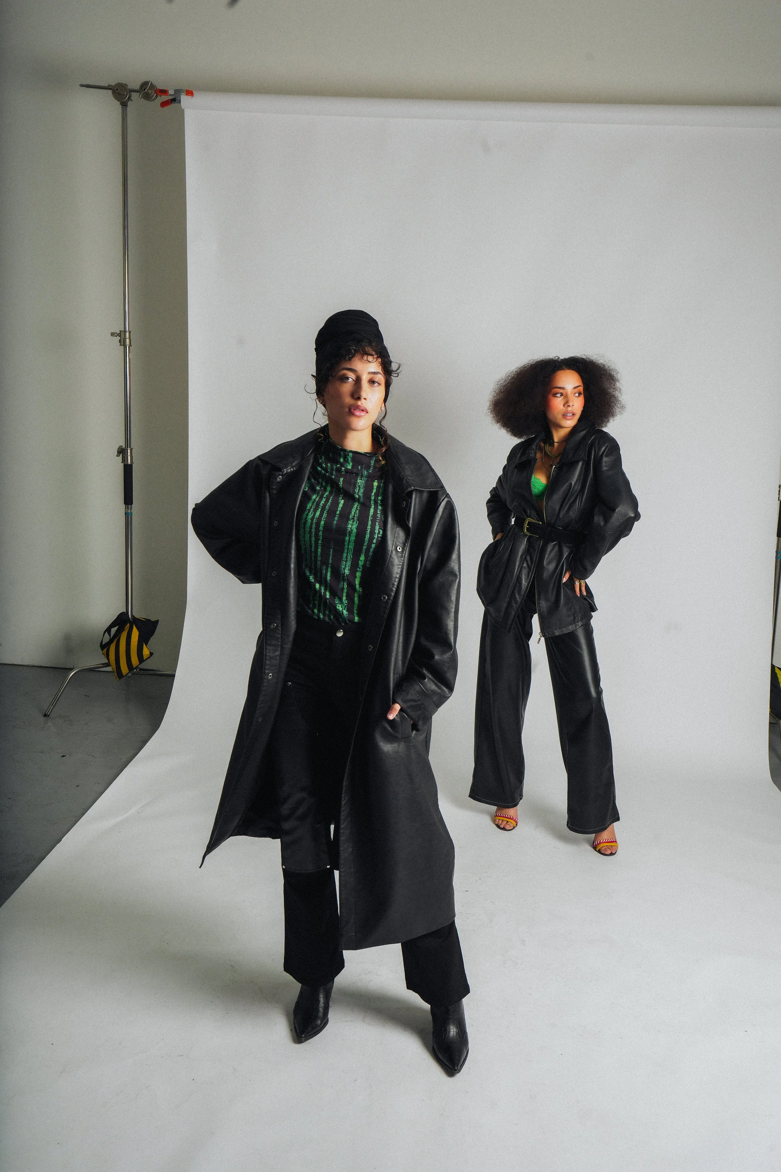 Two models in black leather outfits pose in a photo studio with a white backdrop and studio lighting.