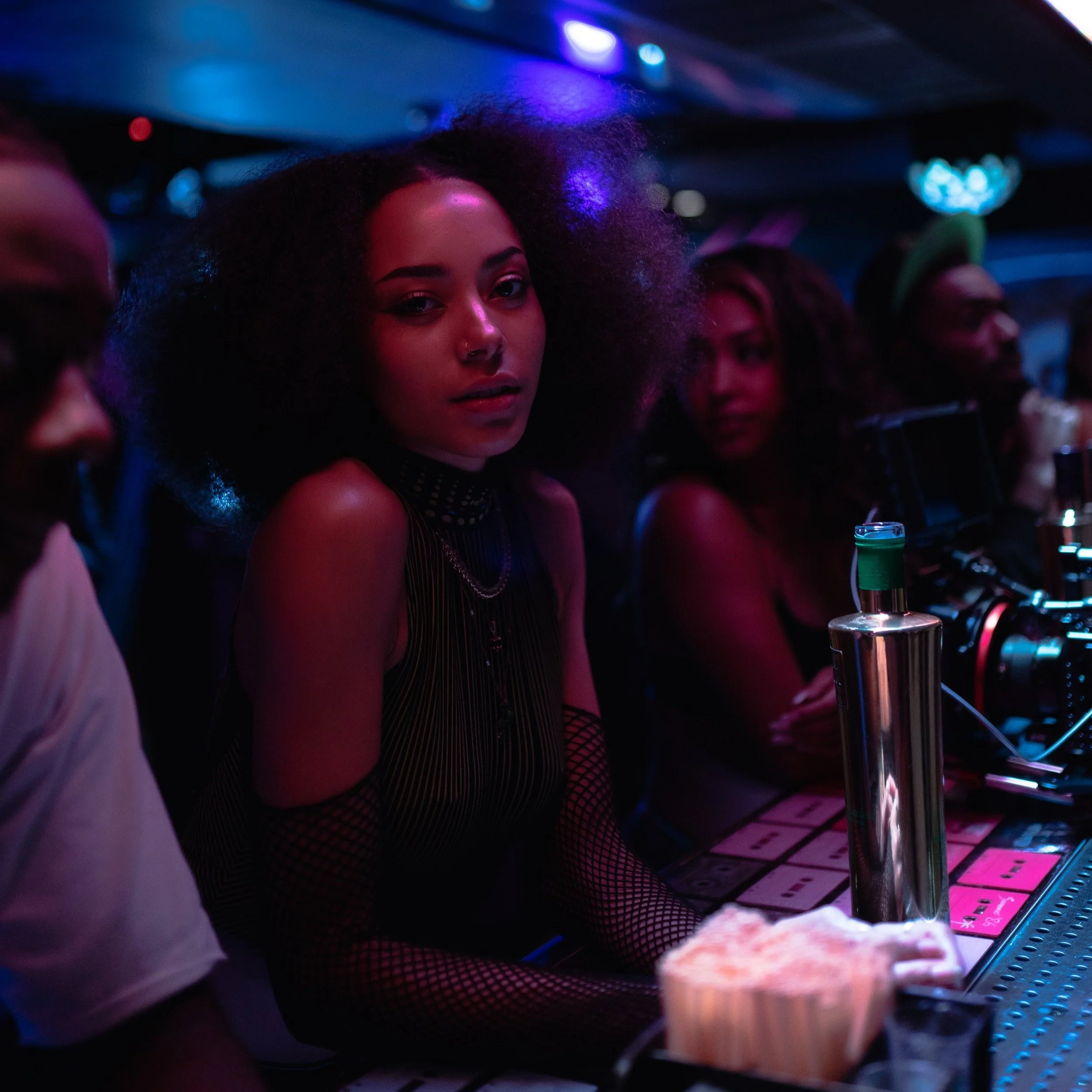 A young woman with curly hair and a nose ring sitting at a table illuminated by colorful blue and purple lights in a dimly lit nightclub or bar, with drinks and a pink menu on the table.