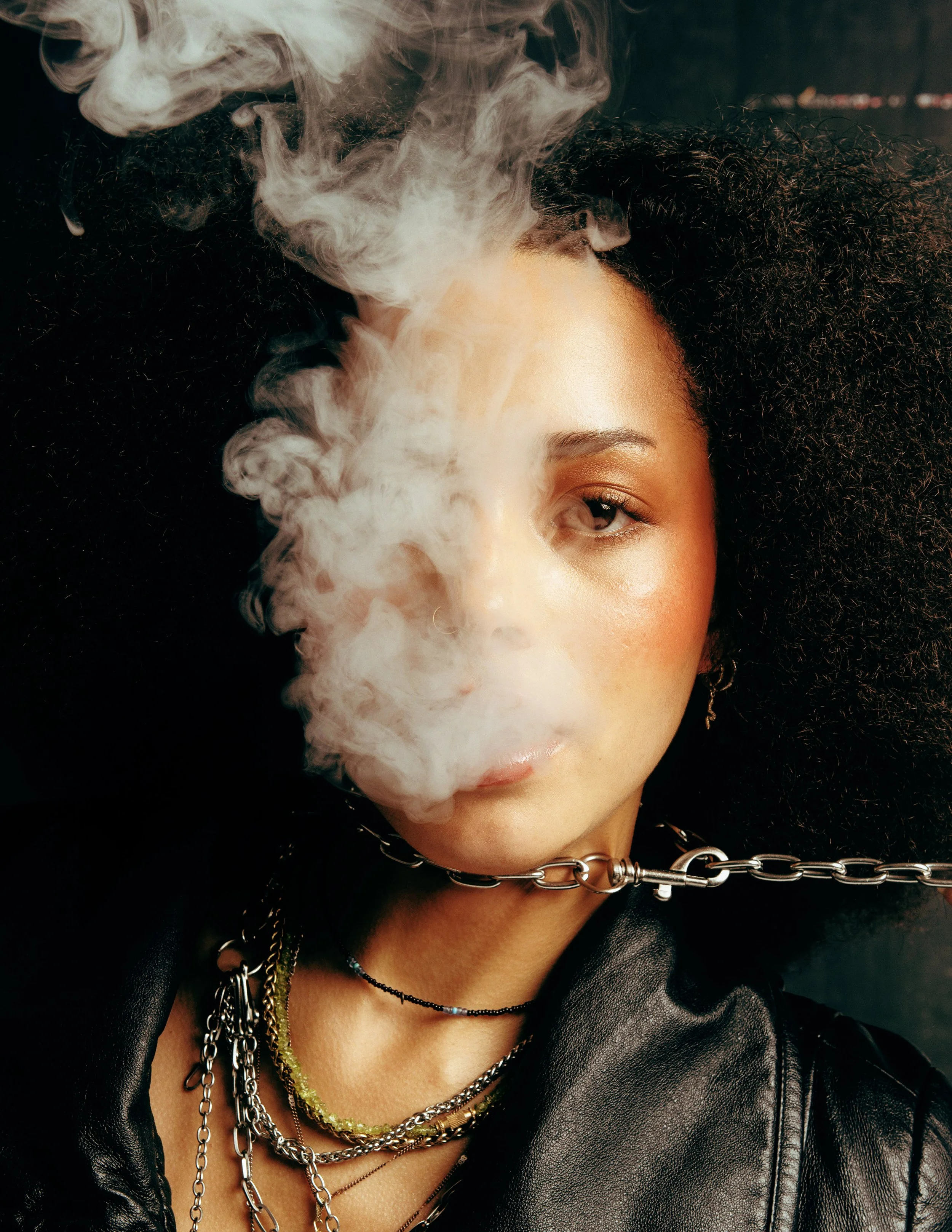 Close-up of a person's face with smoky vapor covering part of one eye, wearing layered chains and a black leather jacket, with curly hair and a chain choker.