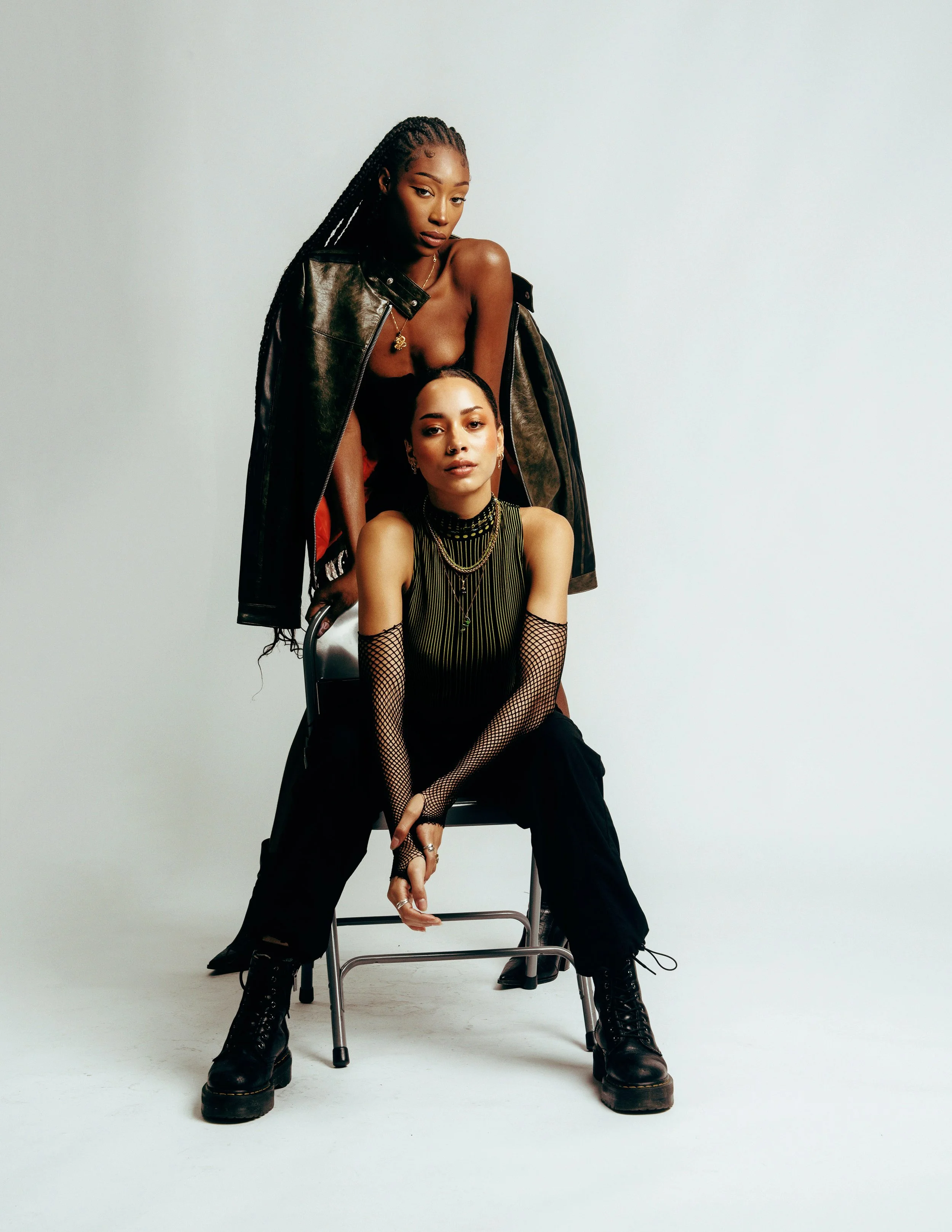 Two young women are posing together against a plain light gray background. One woman, standing, has long, braided hair and is wearing a partially open black leather jacket over a black top, with a gold necklace. The other woman, seated, has short dar