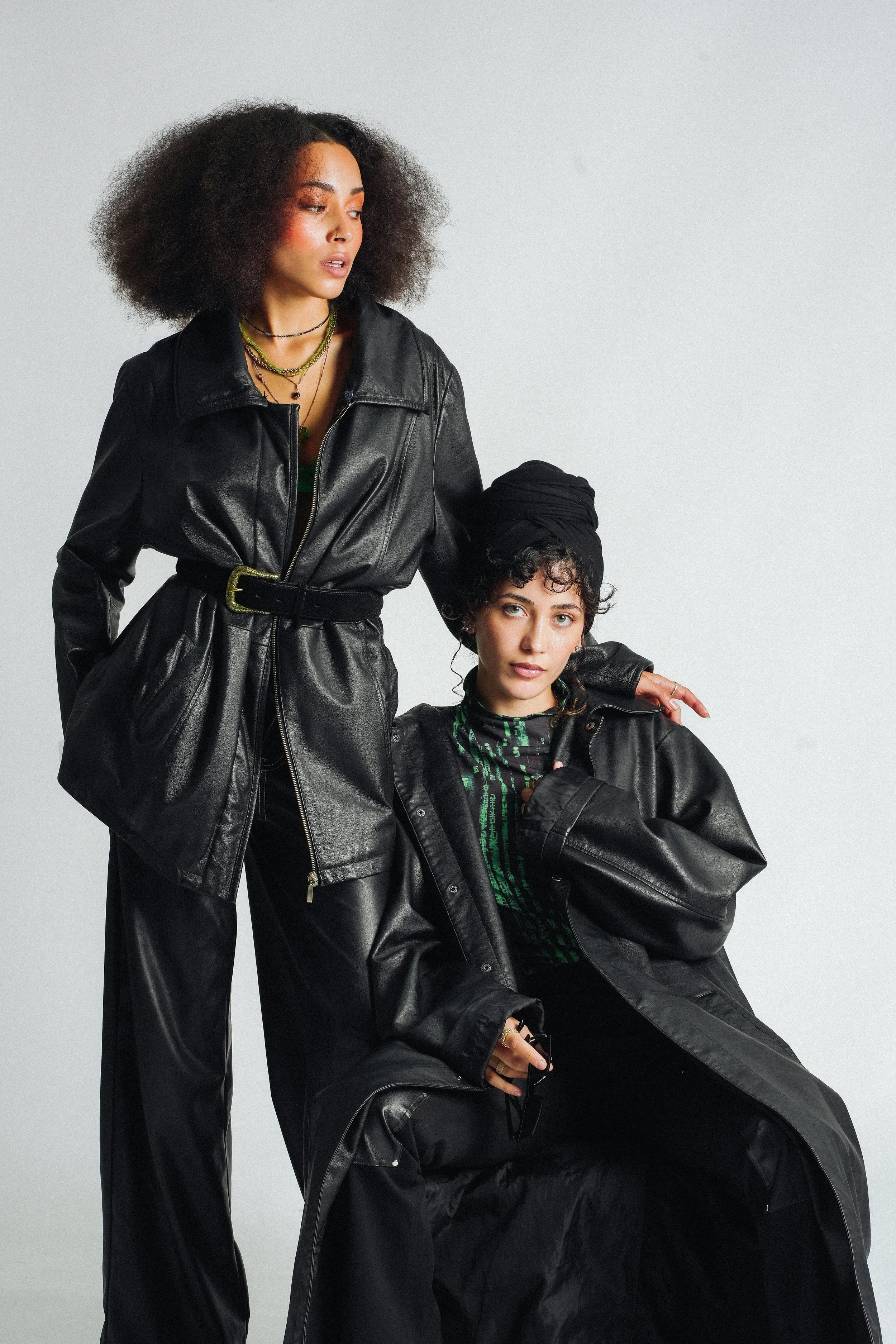Two women with curly hair wearing black leather outfits pose together against a plain gray background.