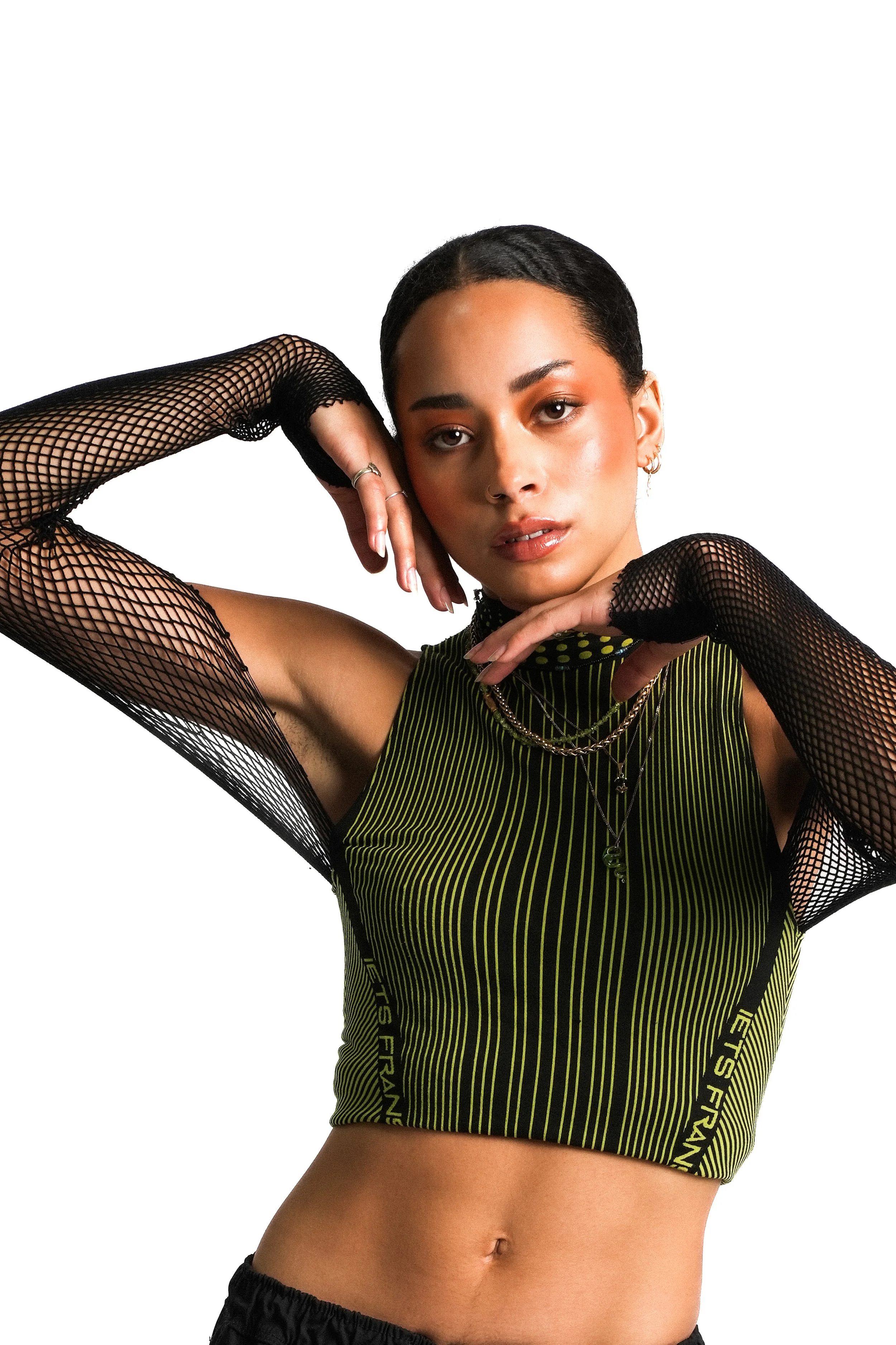 A young woman with short dark hair posing with her arms around her head, wearing a green and black sports tank top and black fishnet gloves, against a white background.